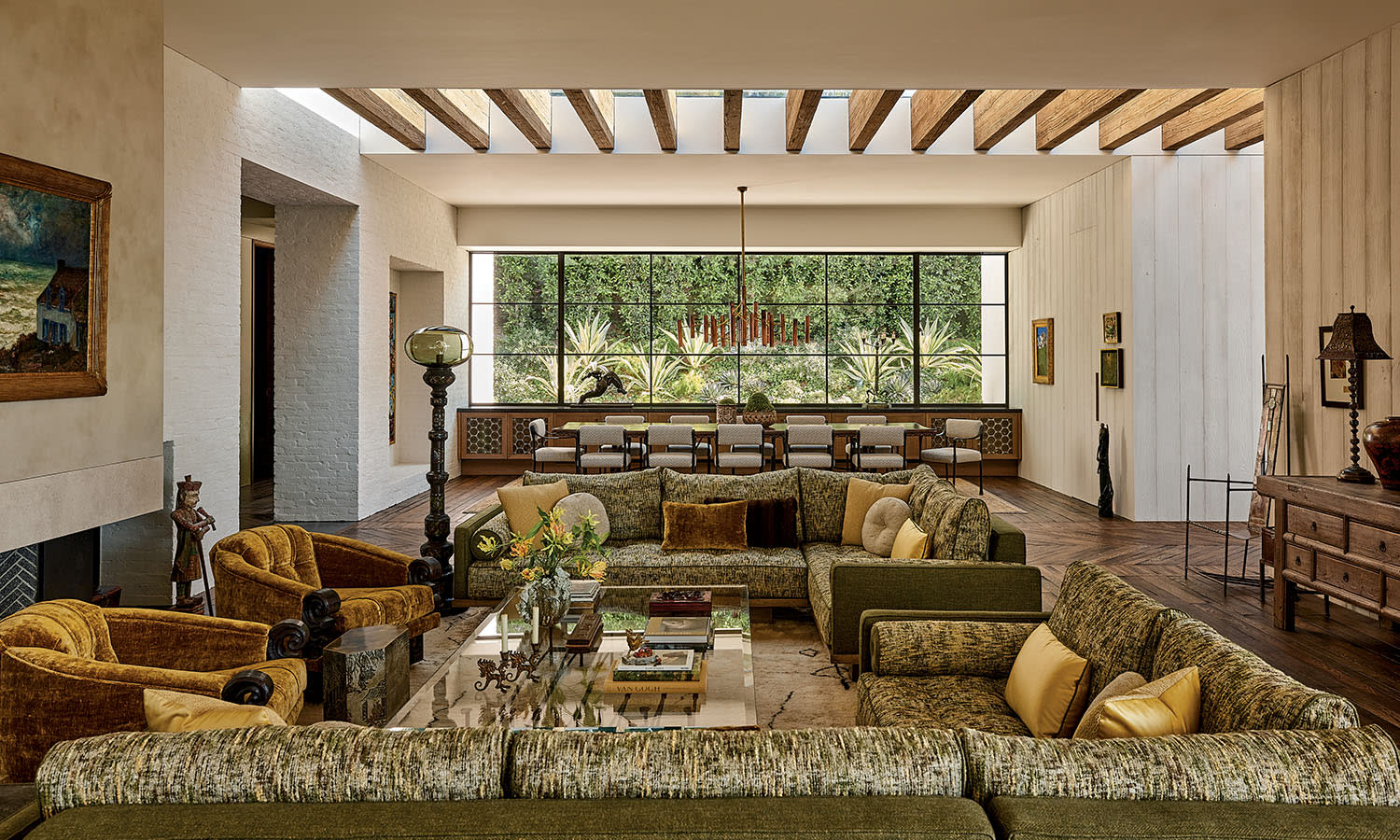 Spacious living room with olive and gold furnishings, beige walls, and a large window revealing a lush garden. Wooden beams and warm lighting create a cozy ambiance.