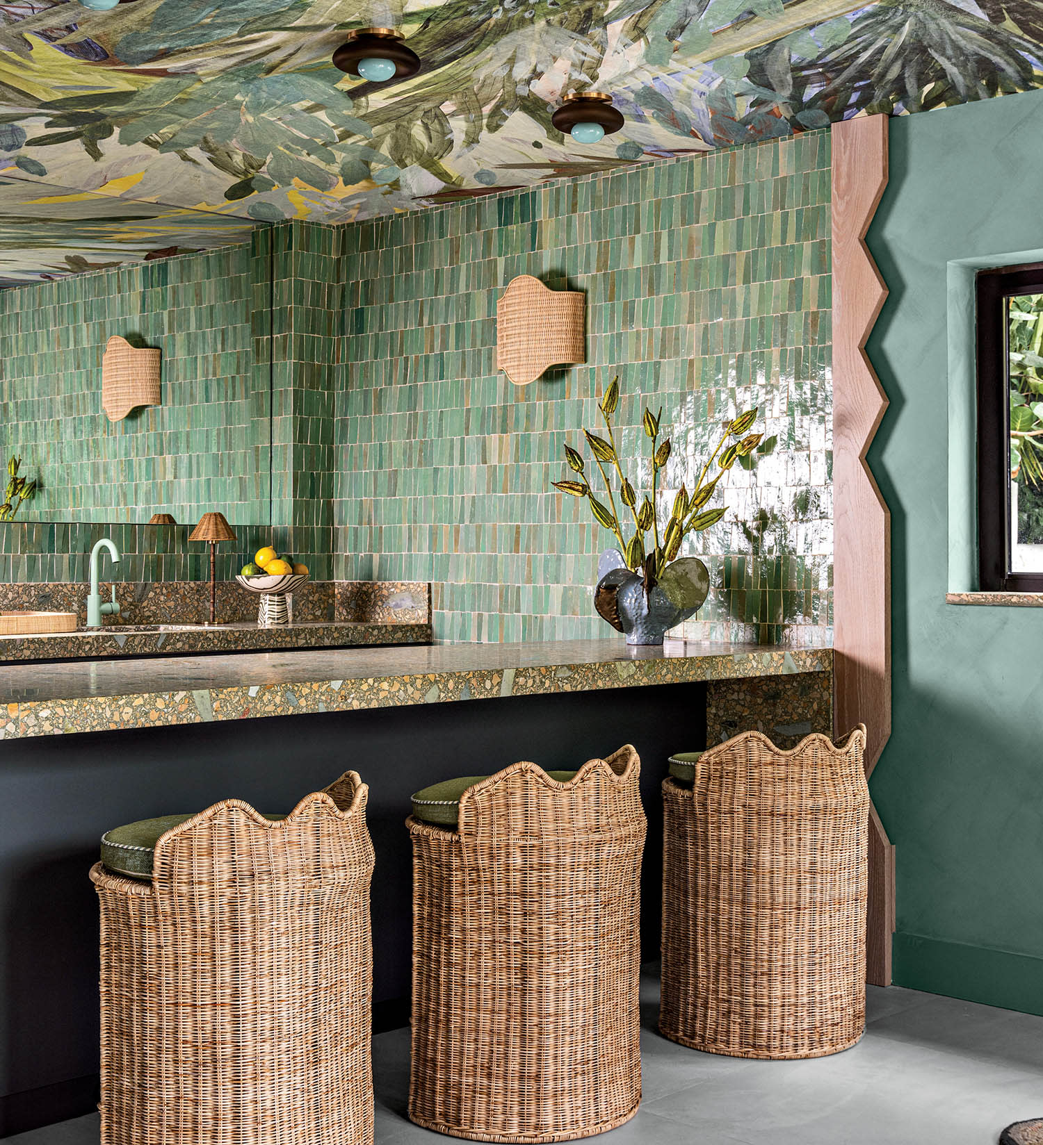 Colorful bathroom with green tiled walls, tropical ceiling mural, wicker stools, and a stone countertop. A vase with flowers adds a touch of nature.