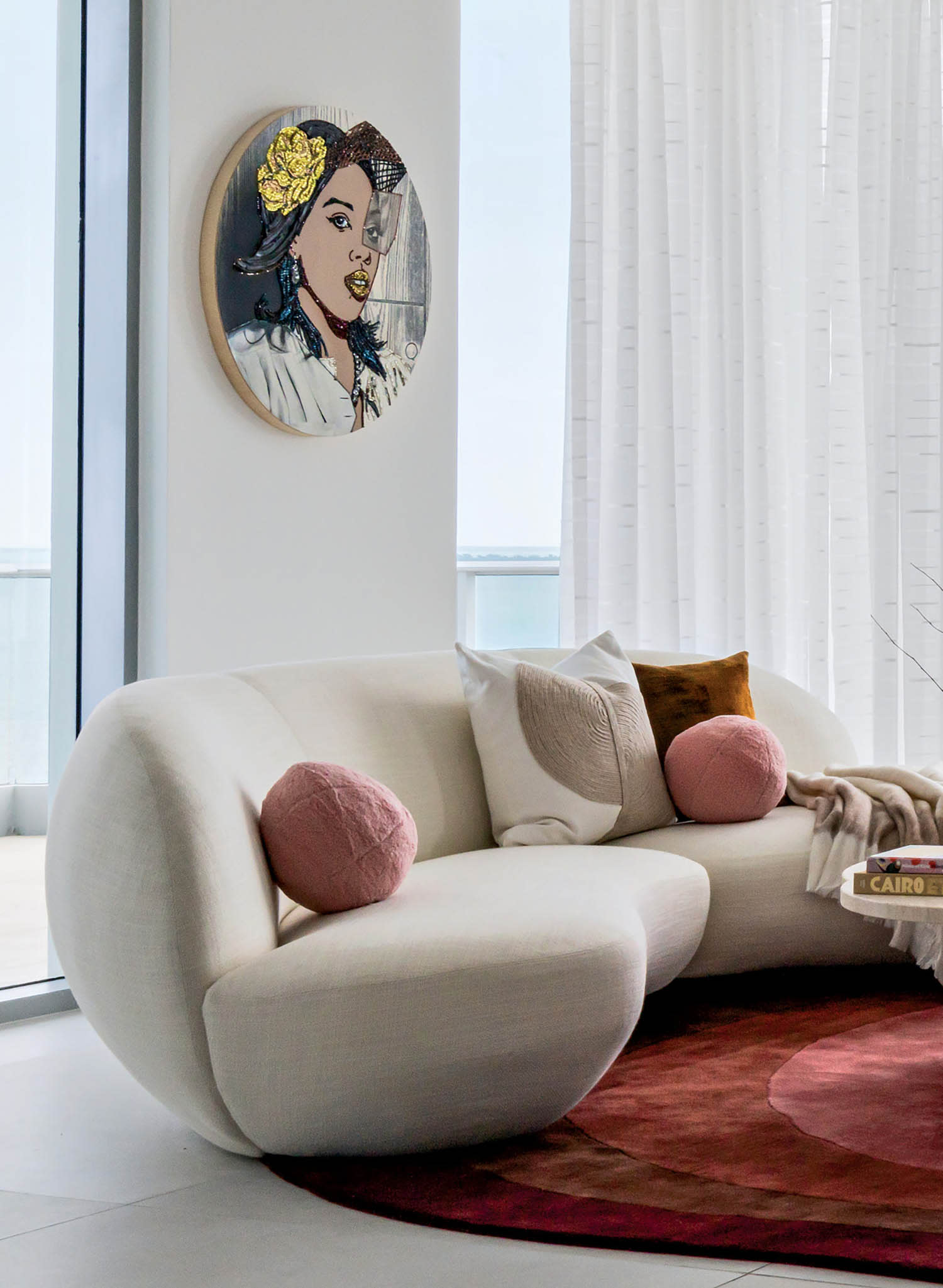 Stylish living room with white curved furniture, round red rug, and large windows. A portrait on the wall and sheer curtains add elegance.