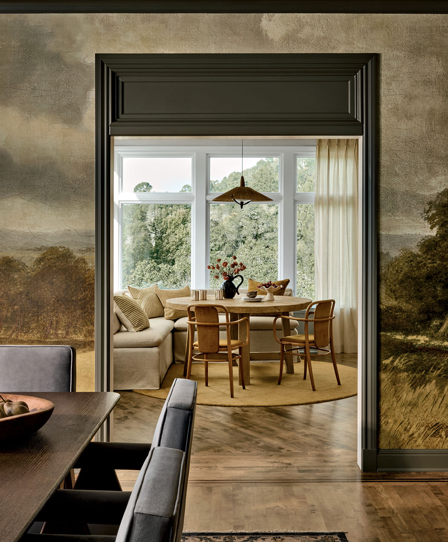 Cozy dining area seen through a doorway, with a round table, wooden chairs, a vase of flowers, cream curtains, and large windows overlooking trees.