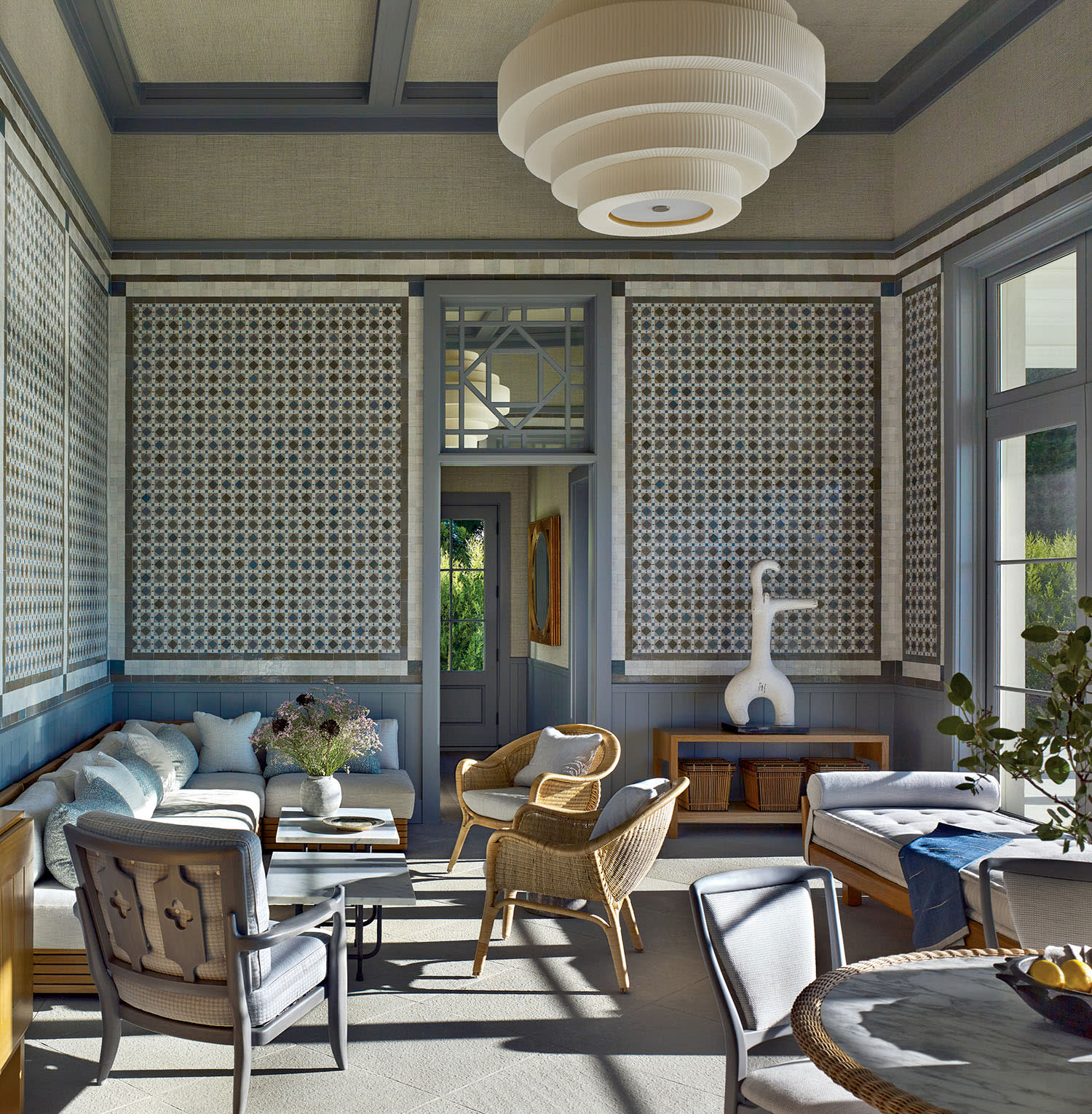 Stylish sunroom with a patterned ceiling and walls, cozy wicker chairs, soft sofas, a central modern table, and a large pendant light. Bright, serene ambiance.