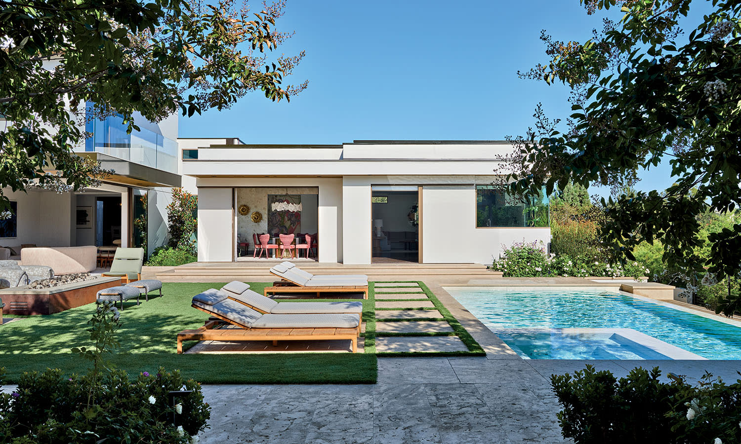 Modern backyard with a pristine pool, sun loungers on manicured grass, and a sleek, white house. Vibrant greenery frames the serene scene.