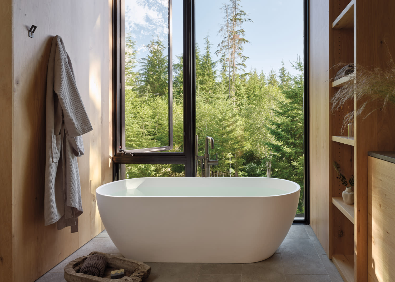 A bathtub in a bathroom overlooks a serene forest view through a large window.