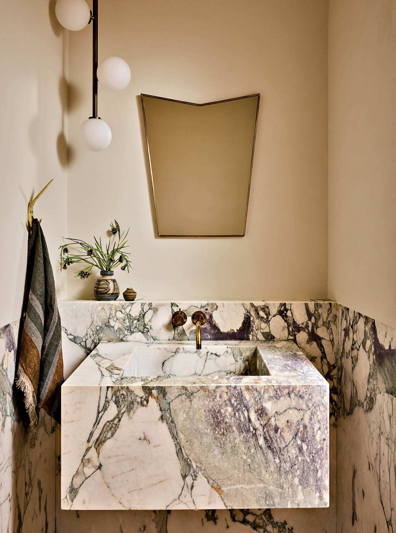 A mirror sits above a marble bathroom sink with a towel hanging on the wall and a plant on the ledge.