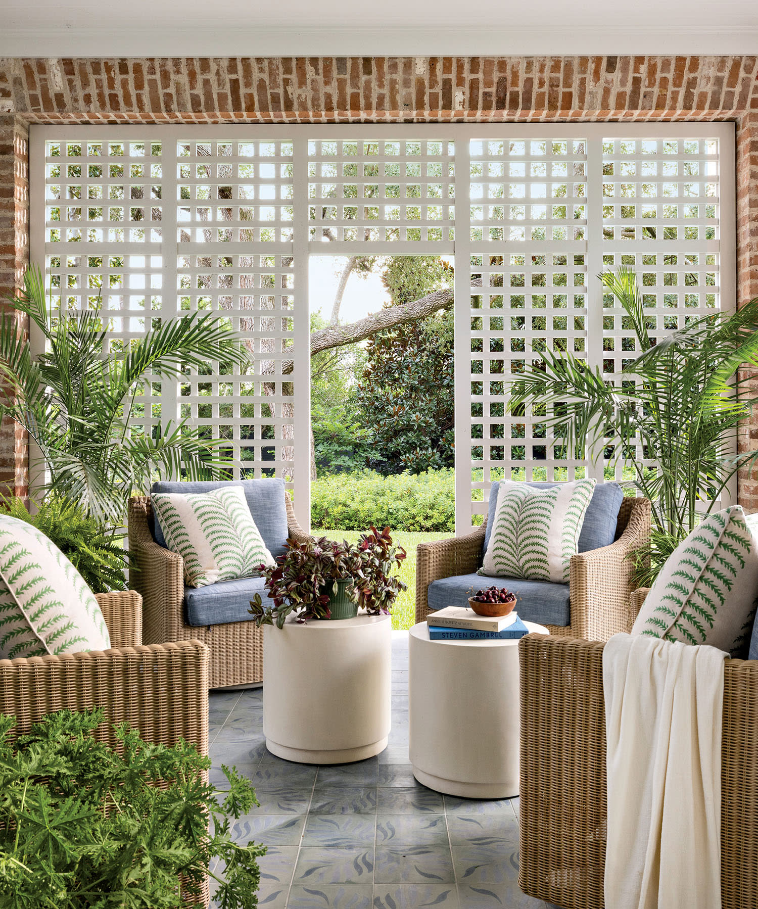 Cozy porch with wicker chairs, blue cushions, and leaf-patterned pillows. White lattice wall, lush greenery outside, and ceiling fan overhead. Calm and inviting.