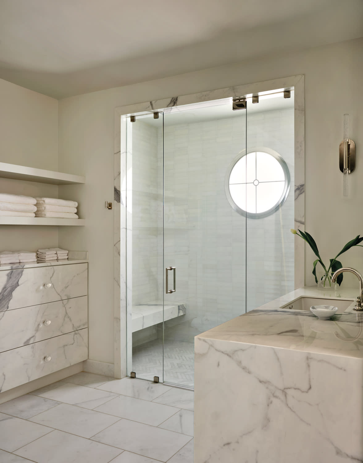 Elegant bathroom with white marble surfaces. Features a glass shower with a round window, neatly folded towels on shelves, and a sleek sink area. Luxurious and serene ambiance.