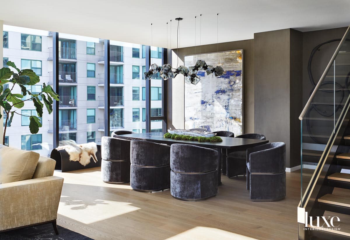 A long black dining table is surrounded by gray-blue velvet chairs. A smoked glass chandelier hangs above the table and a large abstract painting hangs behind it.