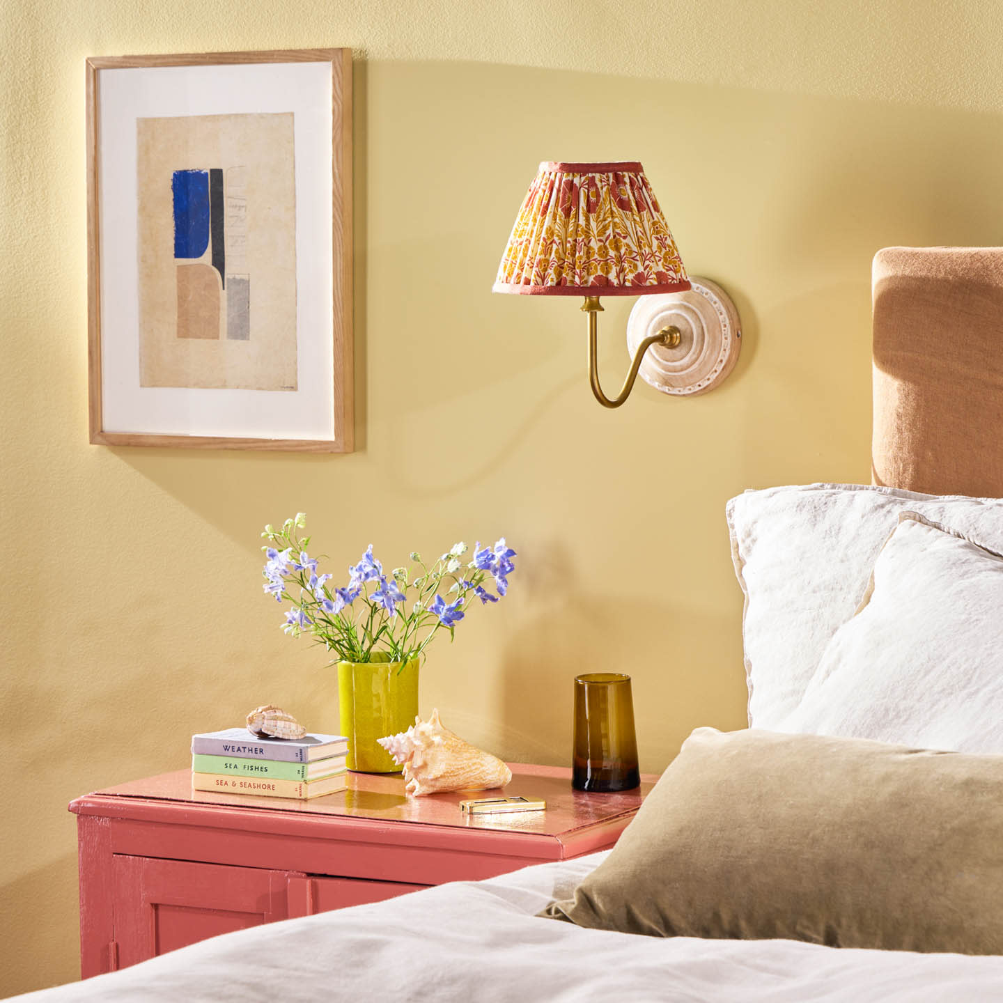 Cozy bedroom scene with a red nightstand. A yellow vase with blue flowers, books, a seashell, and a brown glass sit under a patterned wall sconce. Wall art adds elegance.