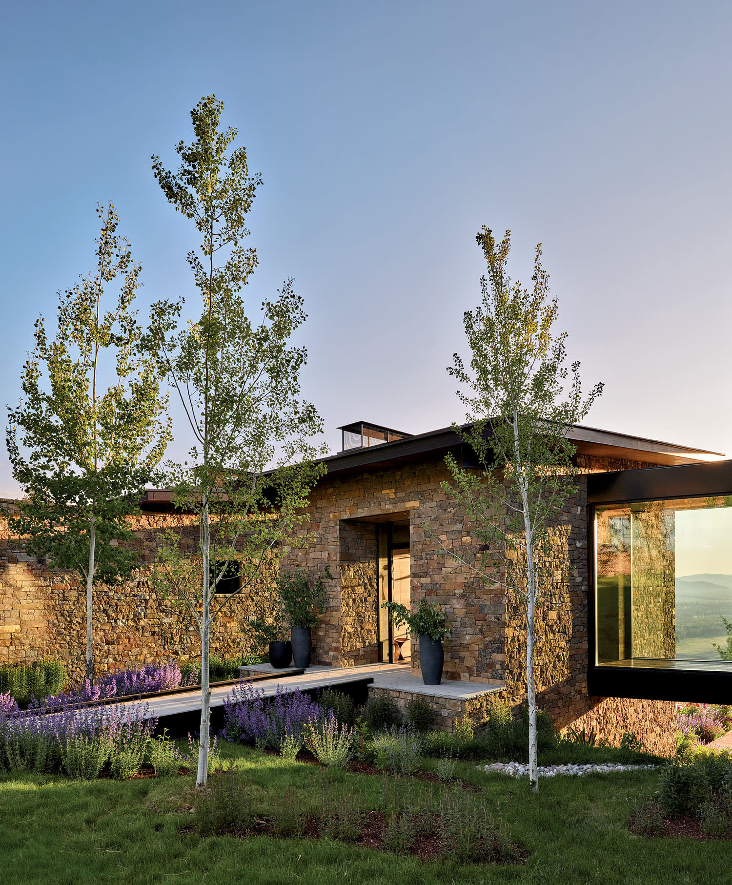 Modern stone house with large windows reflecting a serene landscape, surrounded by lush greenery and young trees under a clear blue sky. Peaceful ambiance.