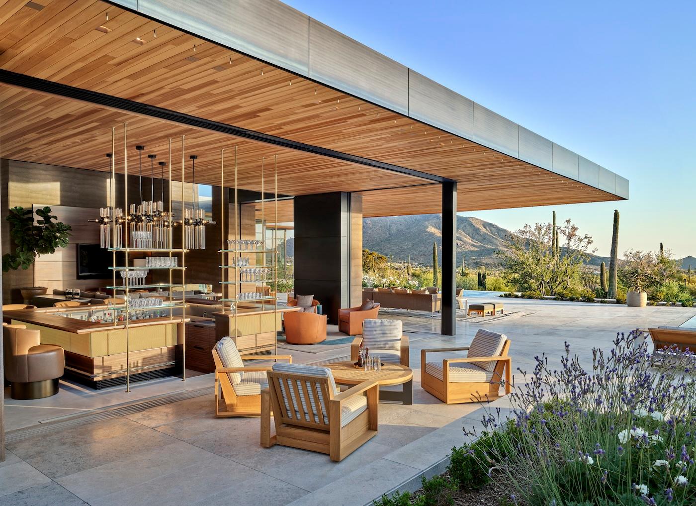 Luxurious outdoor patio with wooden ceiling and furniture. Modern bar area, plush seating, surrounded by desert landscape with mountains and cacti.