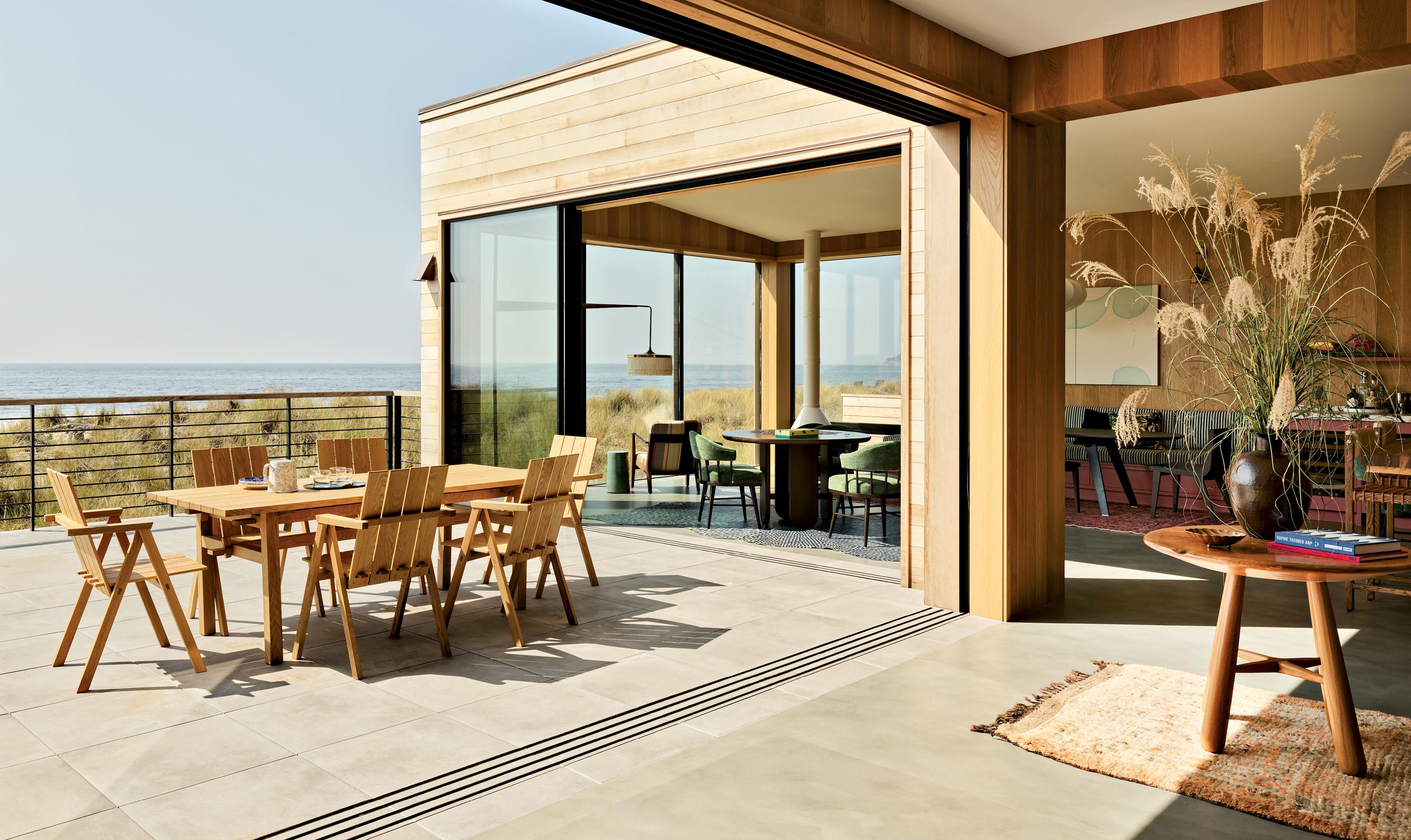 A modern coastal home features an open patio with a wooden dining table set, overlooking the ocean. The interior is warm, with plants and natural decor.