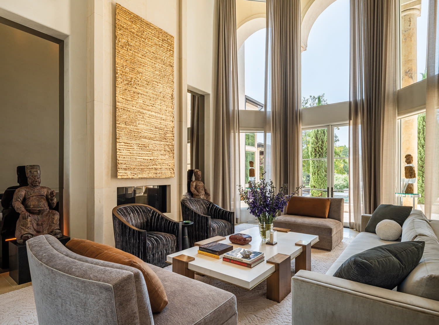 Luxurious living room with tall windows and curtains, featuring modern sofas, a large woven wall art, statues, and a centerpiece table with flowers.