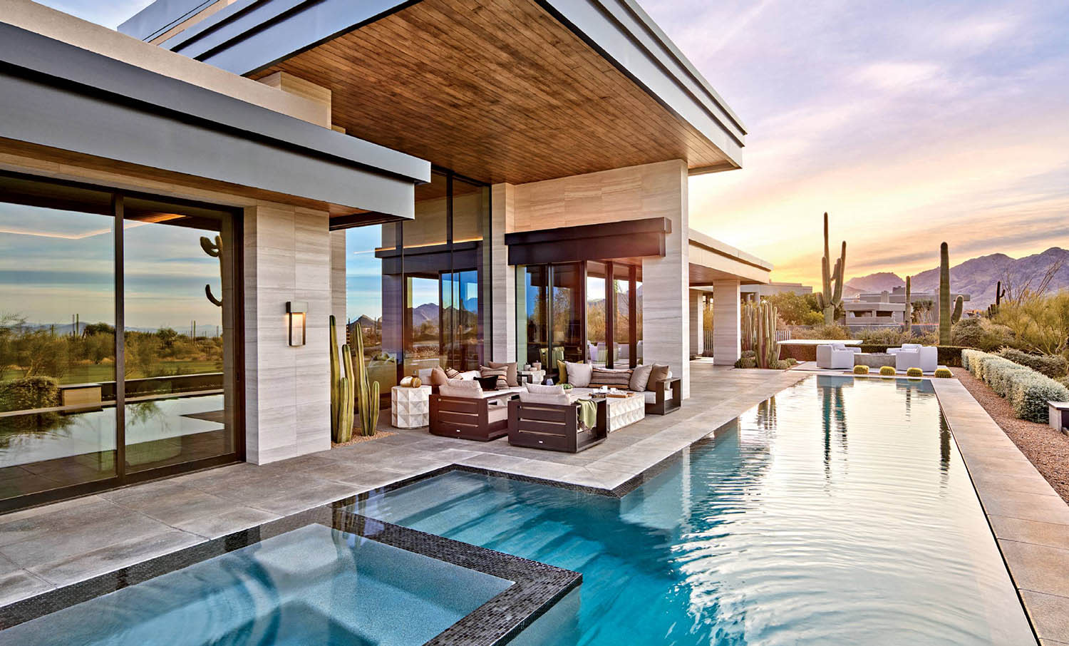 A rectangular swimming pool at the edge of the house, with nearby couches and cacti in the discance.