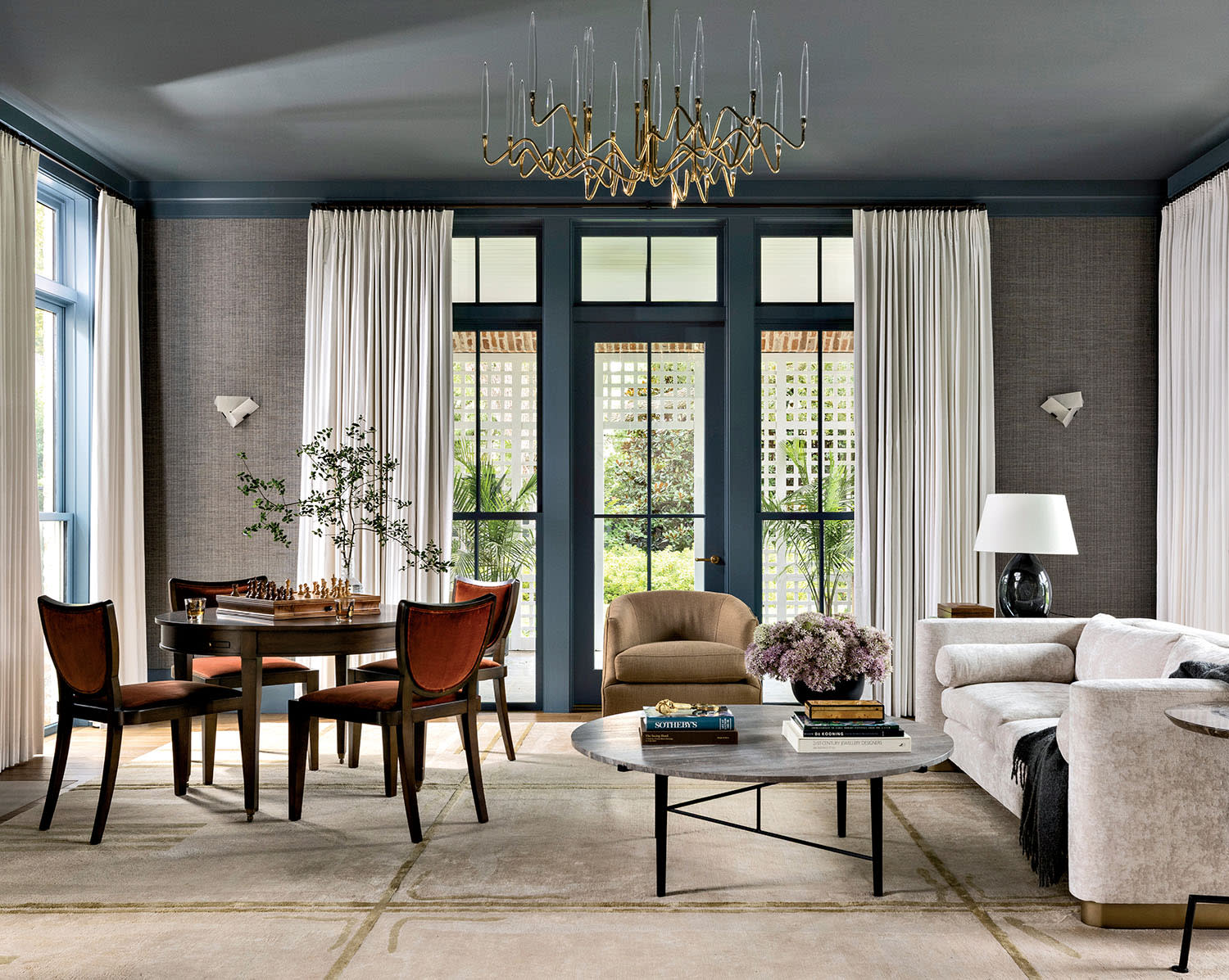 Elegant living room with a modern chandelier, a round chess table with four chairs, and a cozy seating area. Large windows offer a bright, airy feel.