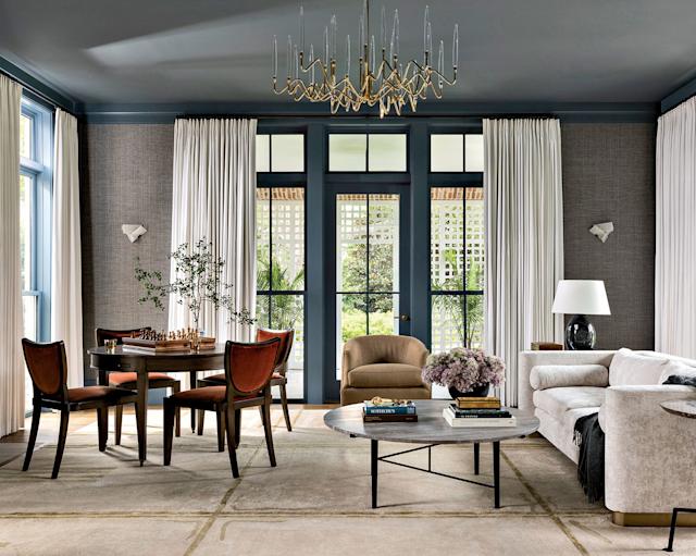 Elegant living room with a modern chandelier, a round chess table with four chairs, and a cozy seating area. Large windows offer a bright, airy feel.