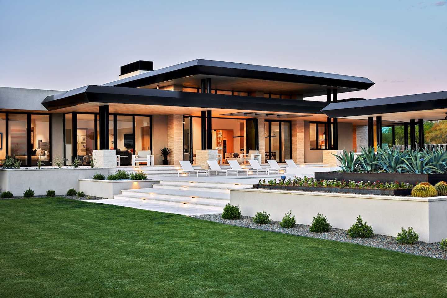 Modern desert-style luxury home at dusk, showcasing bold flat roof overhangs, floor-to-ceiling glass walls, and a tiered limestone patio lined with white chaise lounges. Softly lit steps descend to a manicured lawn framed by sculptural agave, barrel cacti, and neat shrub beds against a pastel twilight sky.