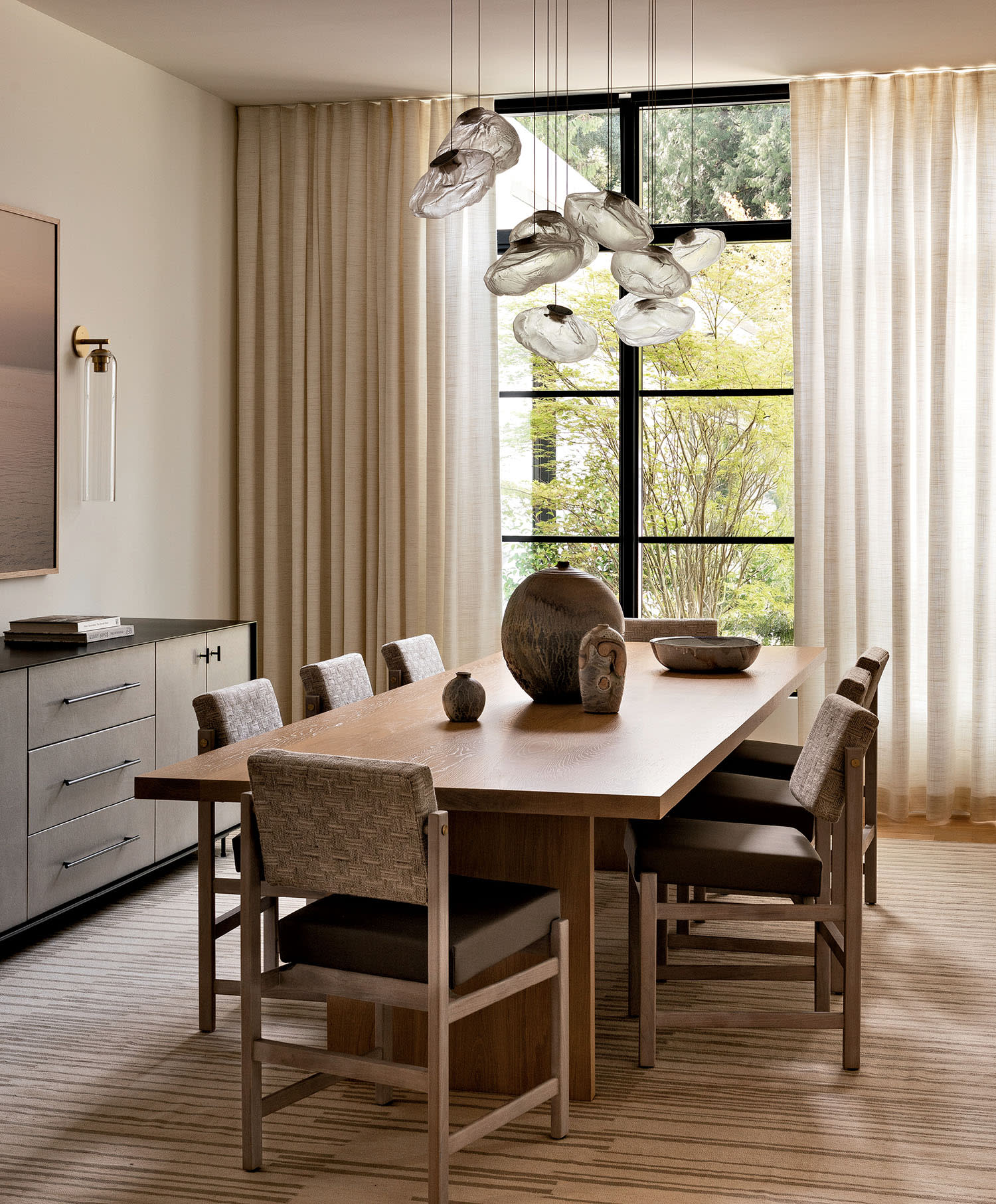 Modern dining room with a large wooden table, eight cushioned chairs, and artistic pendant lights. Neutral tones and tall windows create an elegant ambiance.
