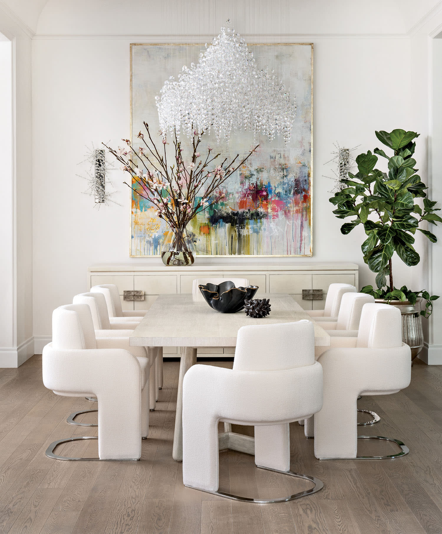 Sophisticated dining room with a modern chandelier, abstract art, and a large table surrounded by white chairs. A vase with branches and a lush plant add elegance.