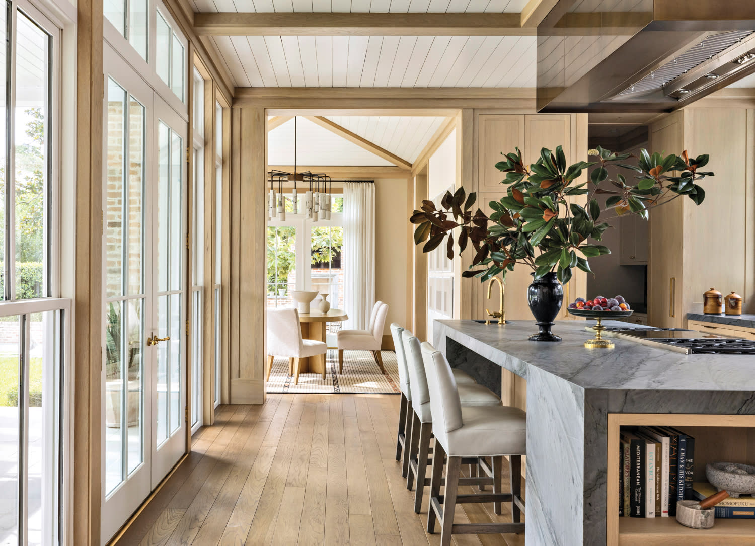 Bright kitchen and dining area with large windows, wooden floors, and light wooden cabinets. A marble island features a vase with green leaves.