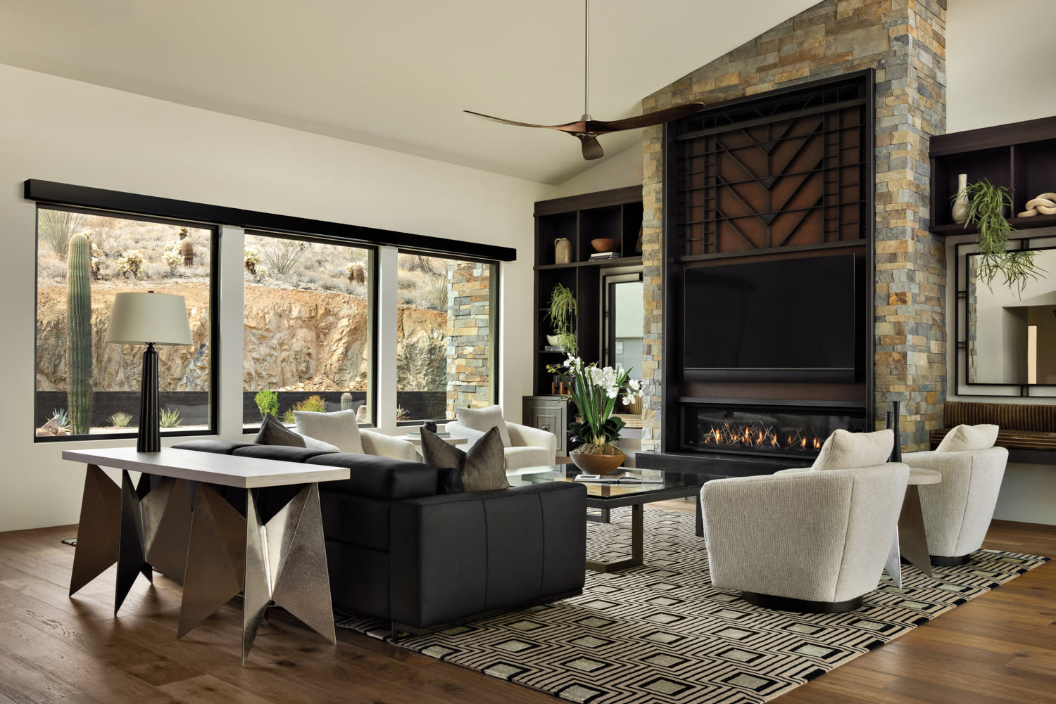 Modern living room with large windows showcasing a desert view. Features include a stone fireplace, black and white furniture, and a geometric rug. Cozy and stylish ambiance.