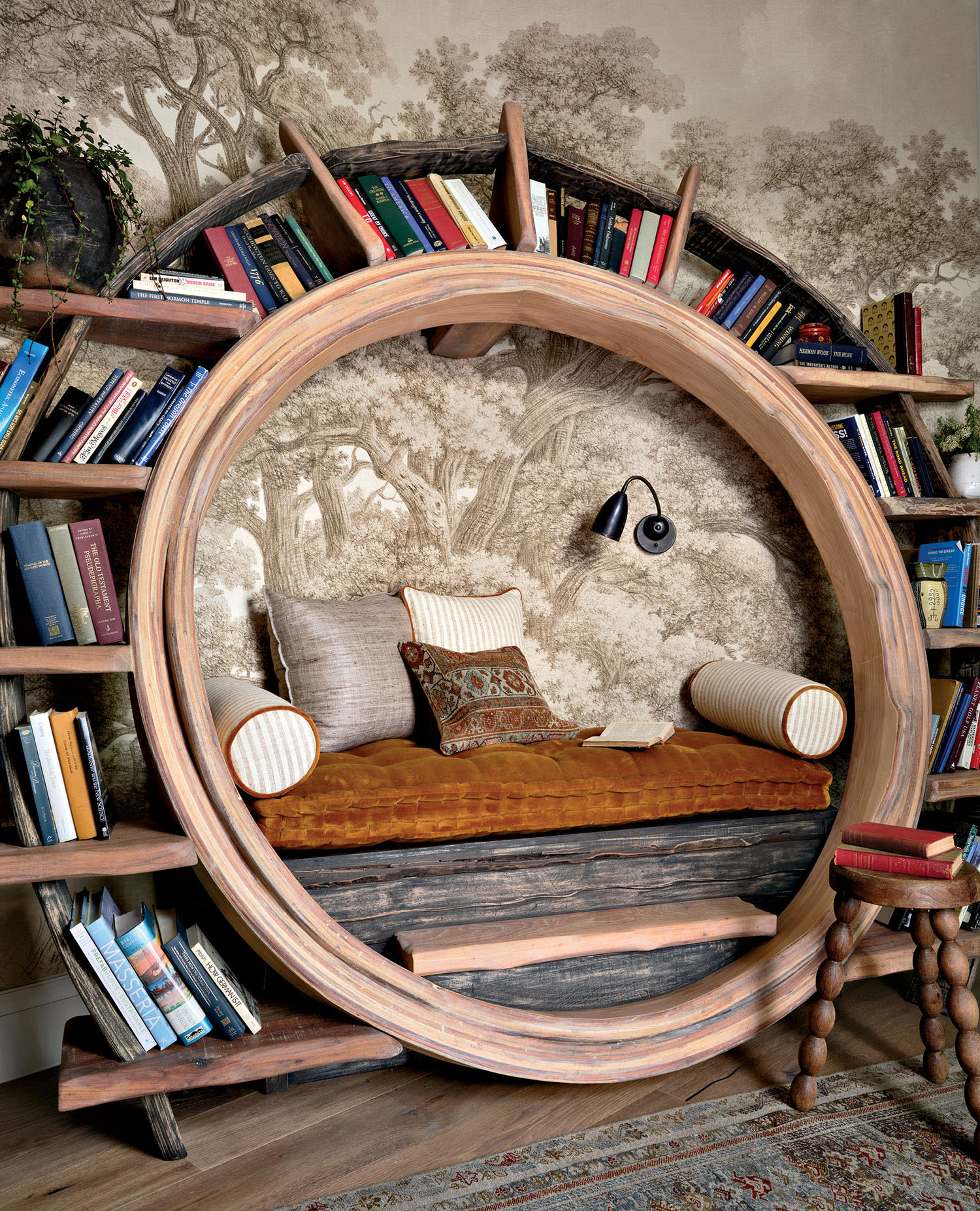 Cozy reading nook inside a circular wooden bookshelf filled with colorful books. The nook has cushions, a plush seat, rustic wallpaper, and a vintage lamp.