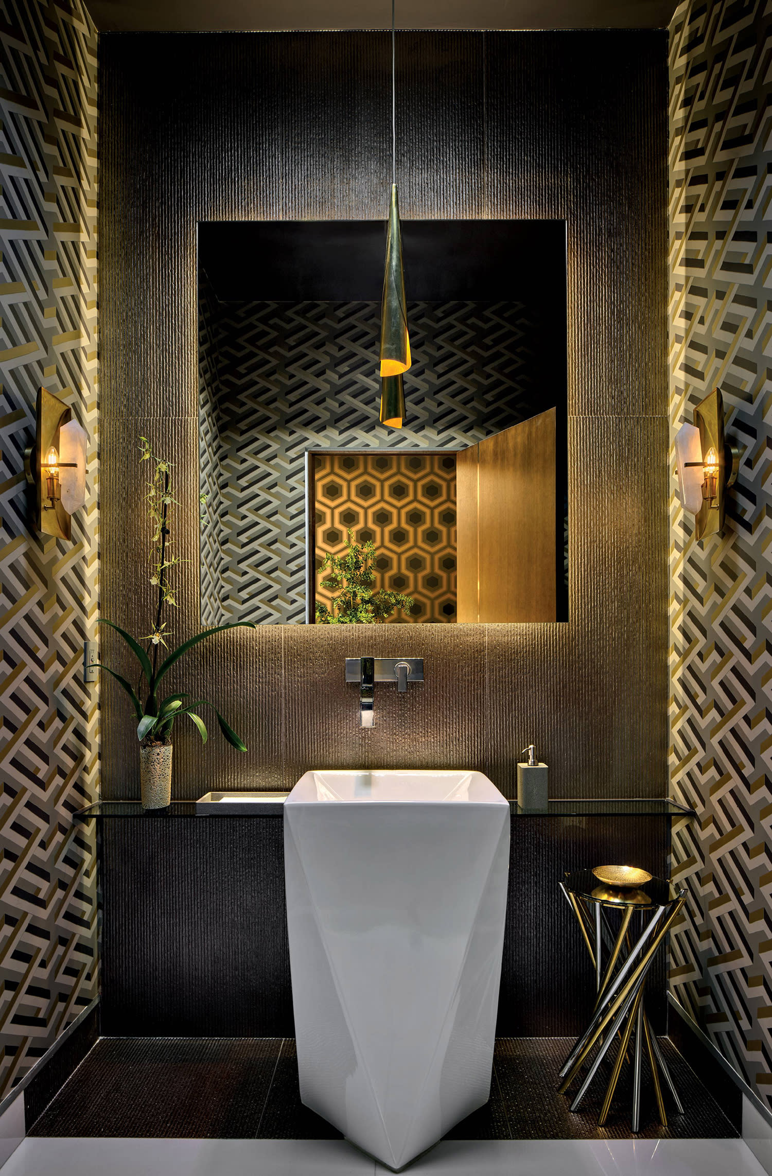 A bathroom decorated with geometric gold wallpaper, a white sink, accent table, and sconce lighting on the walls.