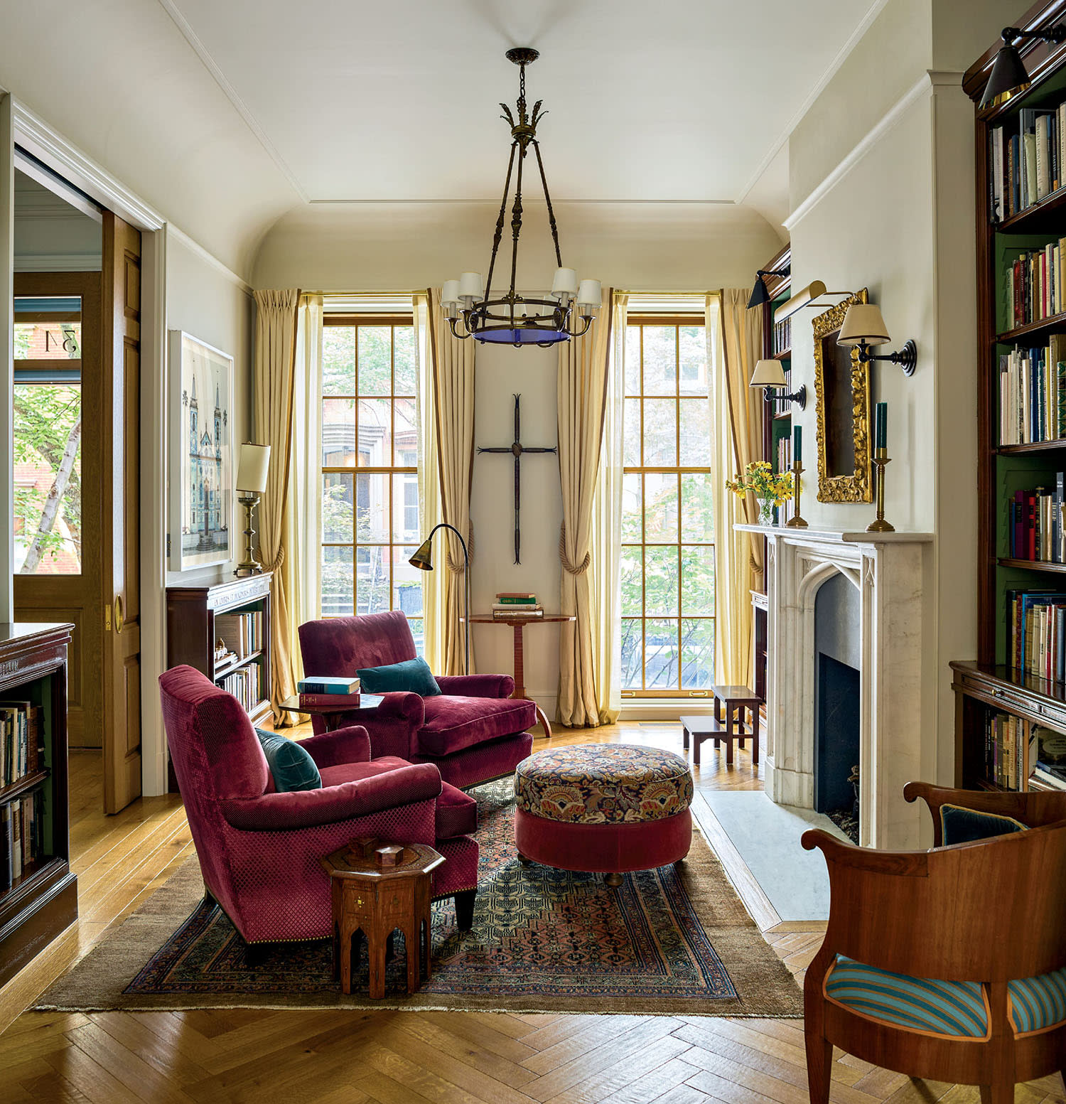 Elegant living room with large windows, burgundy armchairs, and a patterned ottoman on a rug. Floor-to-ceiling bookshelves and a marble fireplace add to the warmth.