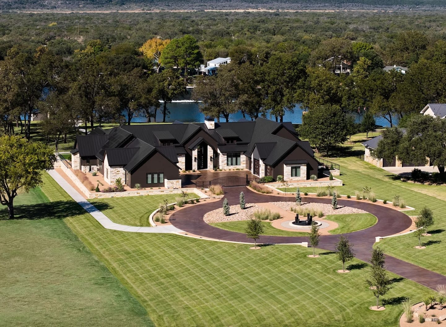 Aerial view of a large, modern home with dark roofs, surrounded by lush green lawns and a circular driveway. Trees and a river are in the background.