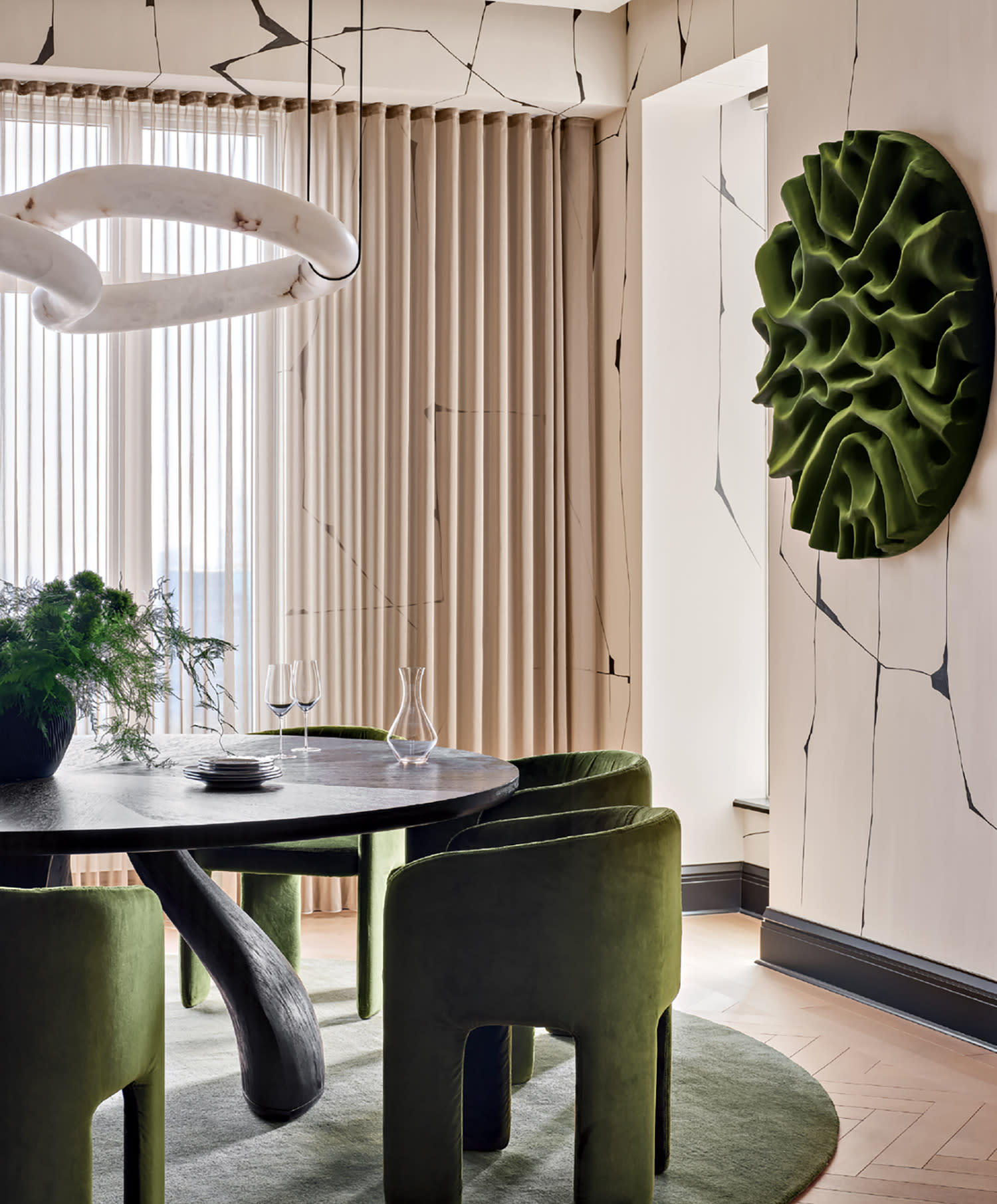 Modern dining room with olive green chairs around a black oval table on a matching green rug. Textured wall art and unique light fixture enhance elegance.