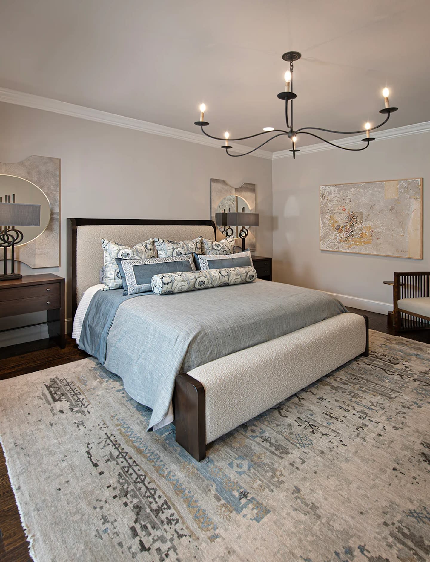 Elegant bedroom with a large bed featuring blue and white patterned pillows, a modern chandelier, abstract wall art, wooden furniture, and a textured rug.