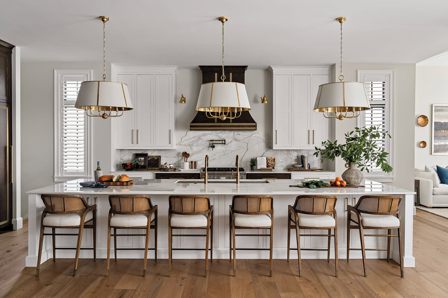 Stylish kitchen with a large white island, six wicker chairs, marble backsplash, brass fixtures, and two elegant chandeliers