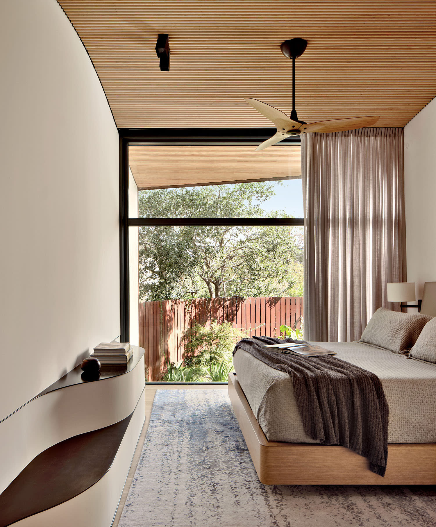 Modern bedroom with a wooden ceiling and large floor-to-ceiling window. Minimalistic design features neutral tones, a double bed, and outside view of trees.