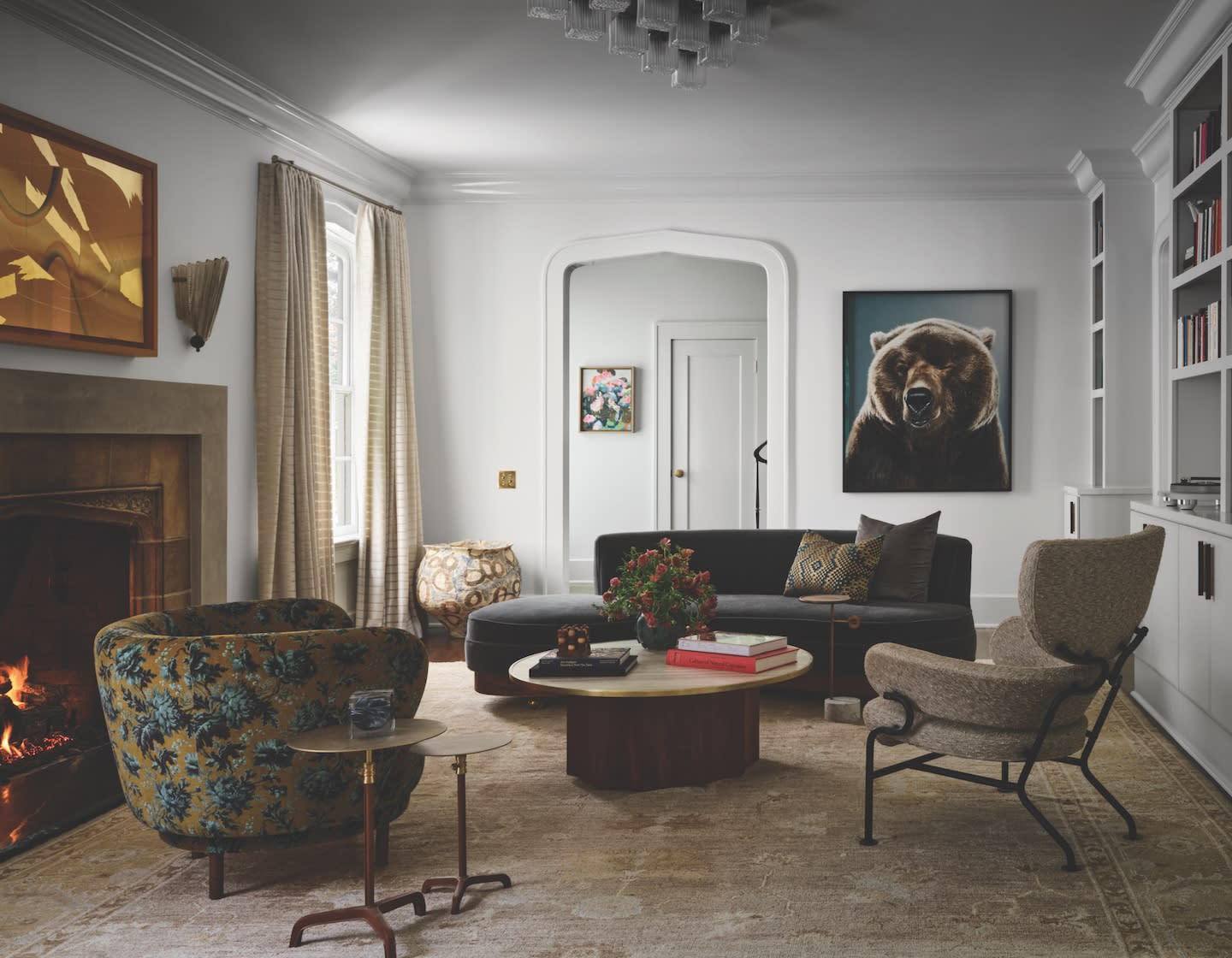 Sophisticated living room with eclectic decor. Features a cozy fireplace, a floral armchair, dark sofa, bear art, and a modern chandelier. Elegant and warm ambiance.