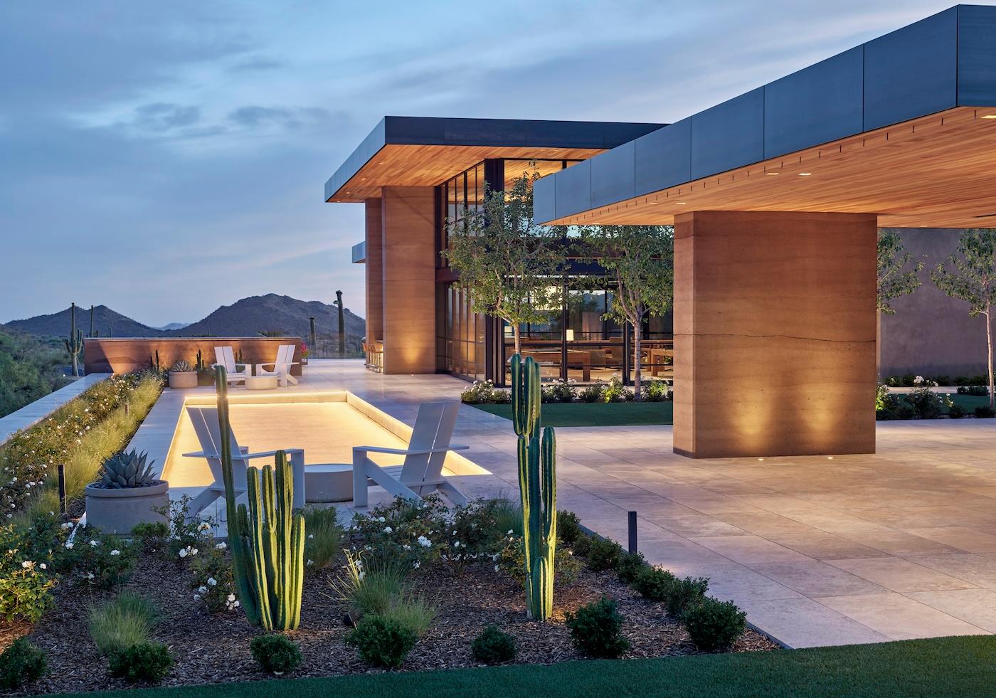 Modern home at dusk with sleek design, illuminated pool, and adjacent patio featuring chairs. Desert landscape with cacti, mountains, and soft lighting.