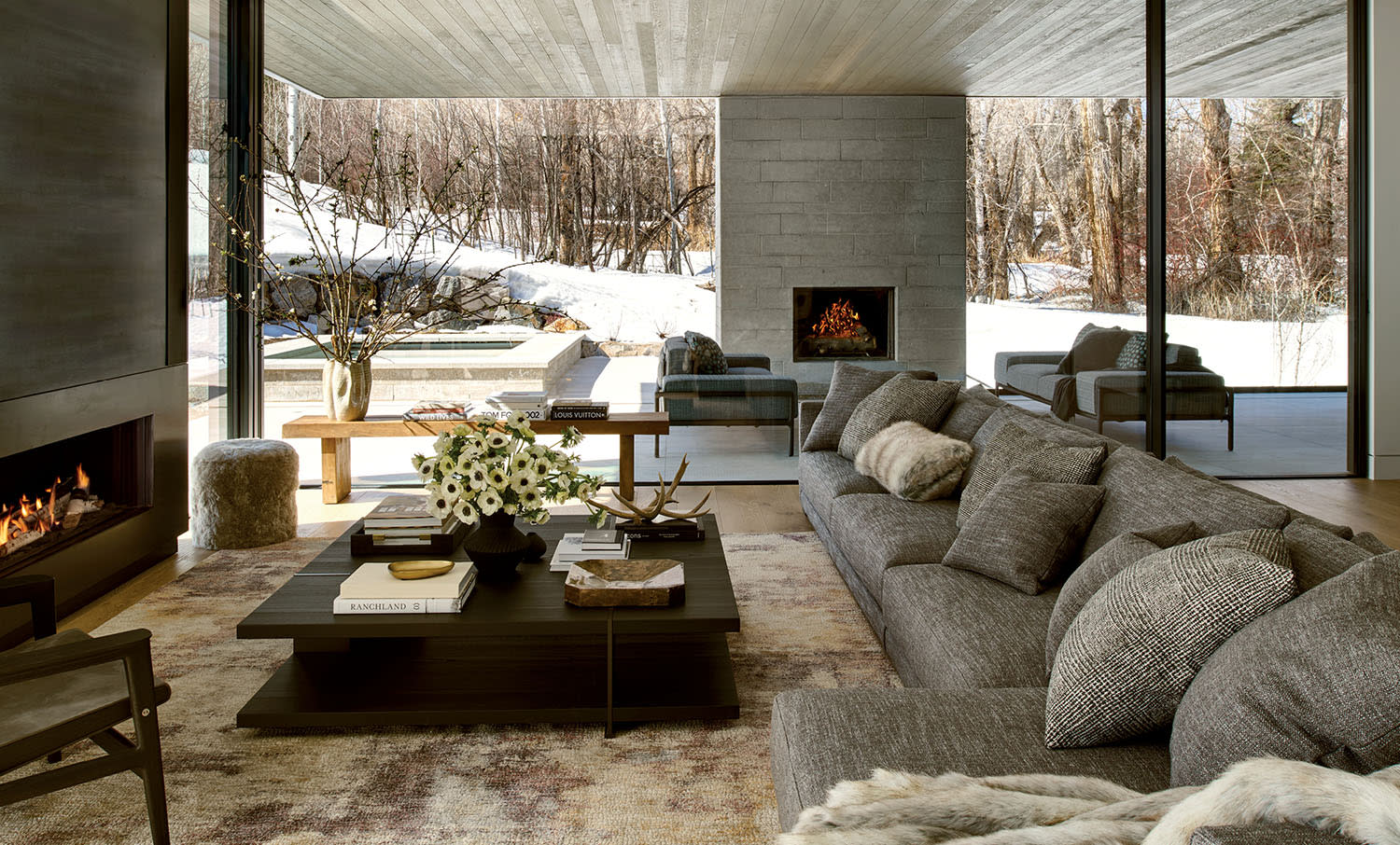 Modern living room with a large gray sofa and cushions, a dark coffee table with flowers, a fireplace, and a glass wall overlooking snowy woods.