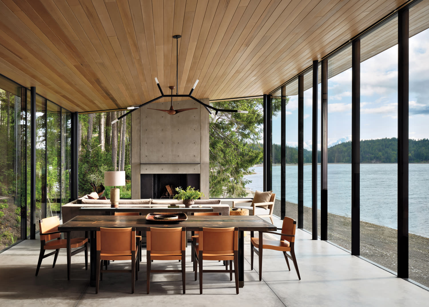 Modern dining room with floor-to-ceiling windows, overlooking a serene lake. Features a wooden table, leather chairs, and a concrete fireplace. Airy and tranquil.
