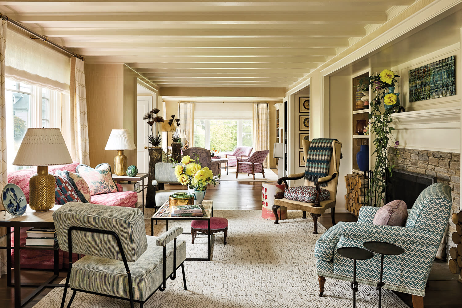 Elegant living room with patterned armchairs and sofas, warm lighting, and a stone fireplace. Bright flowers add a lively touch, creating a cozy ambiance.
