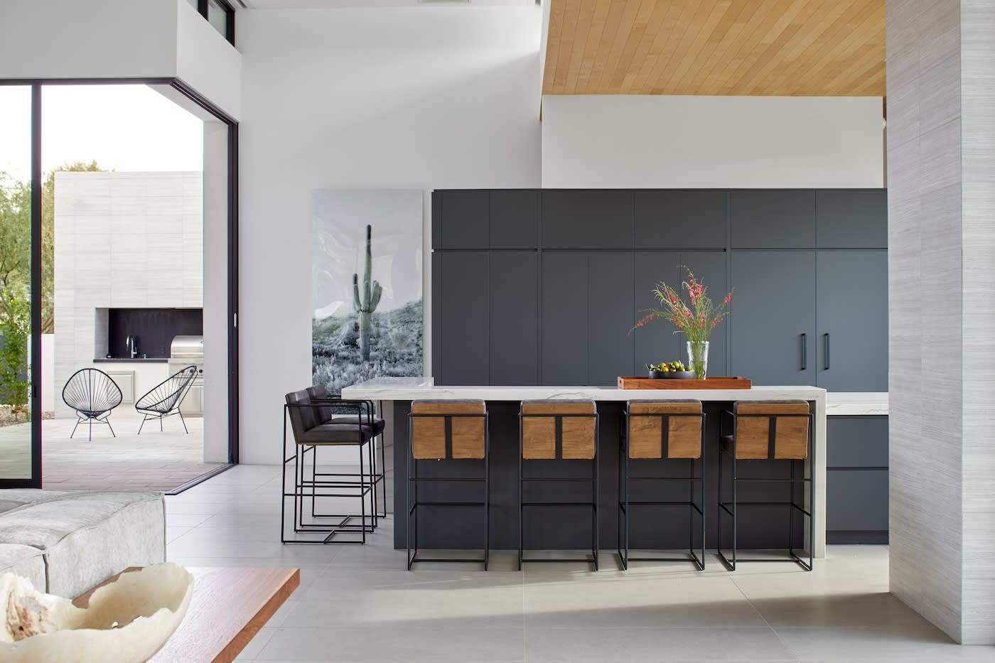 Modern kitchen with sleek black cabinets and wooden accents. A large island with stools faces a spacious patio area. Bright, minimalist, and inviting.