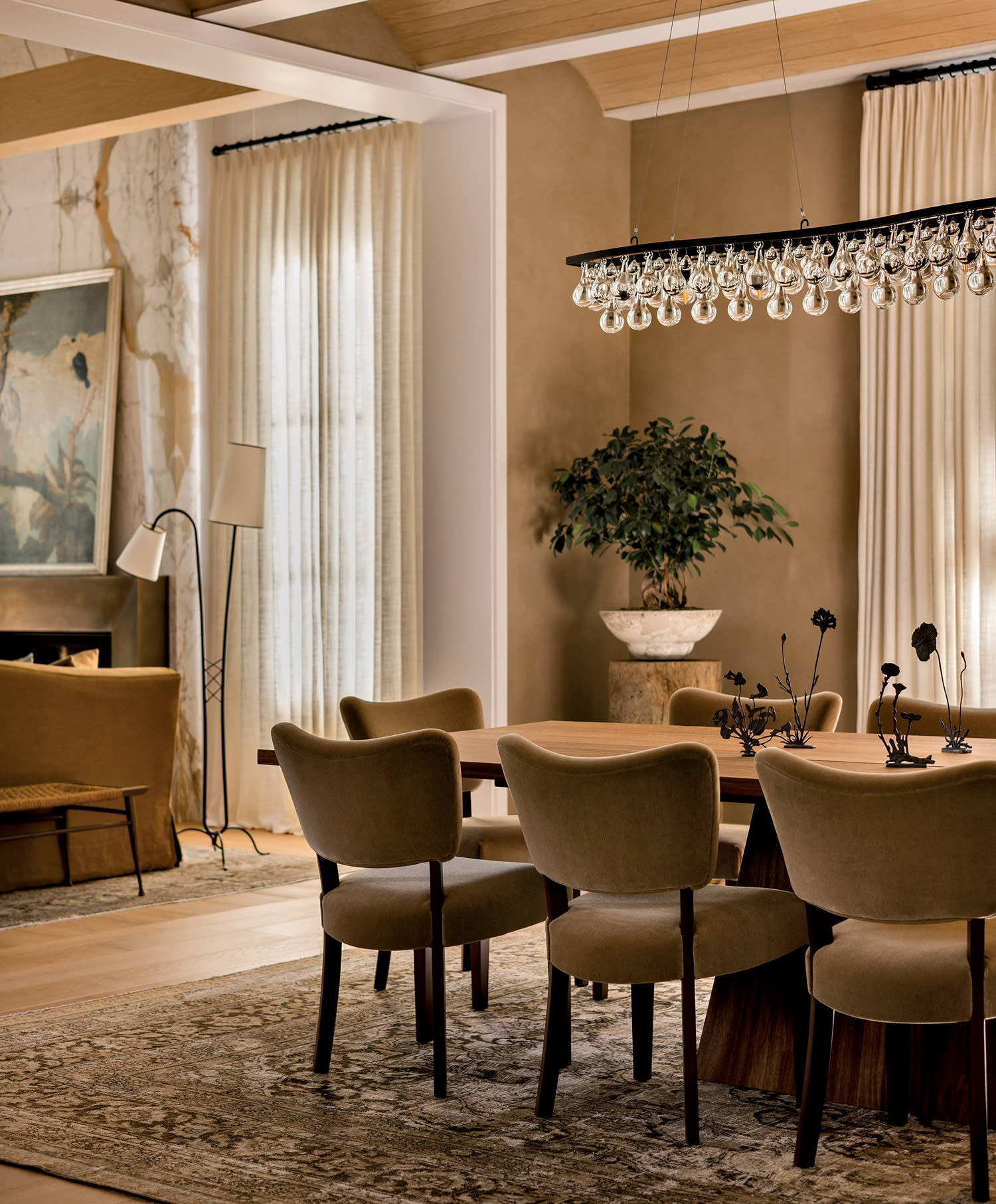 A dining room table and chairs sits on a floral rug with a chandelier above and a plant in the corner.