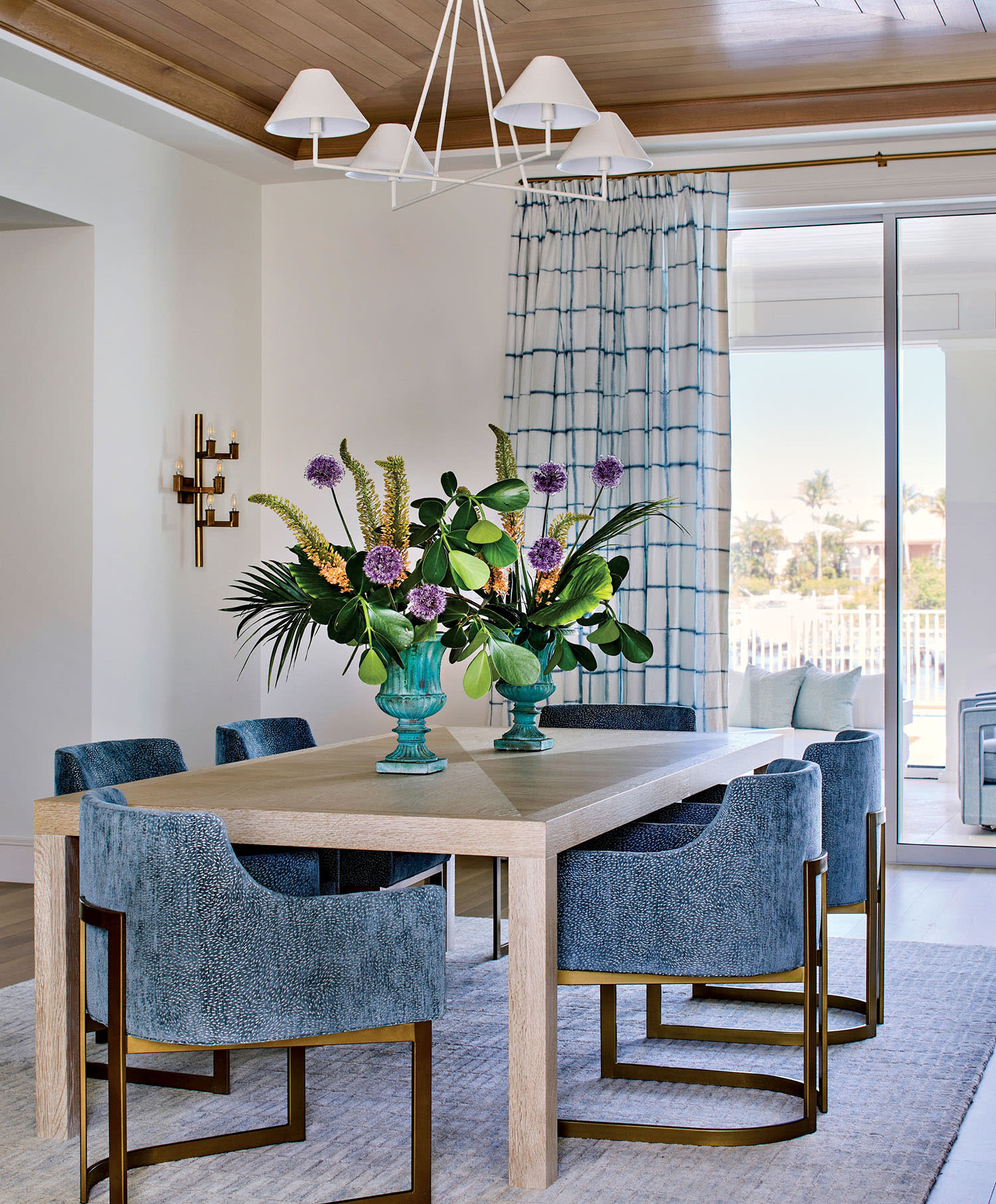 Elegant dining room with a light wood table, blue textured chairs, and vibrant floral centerpiece. Blue-check curtains and large windows. Bright, airy feel.
