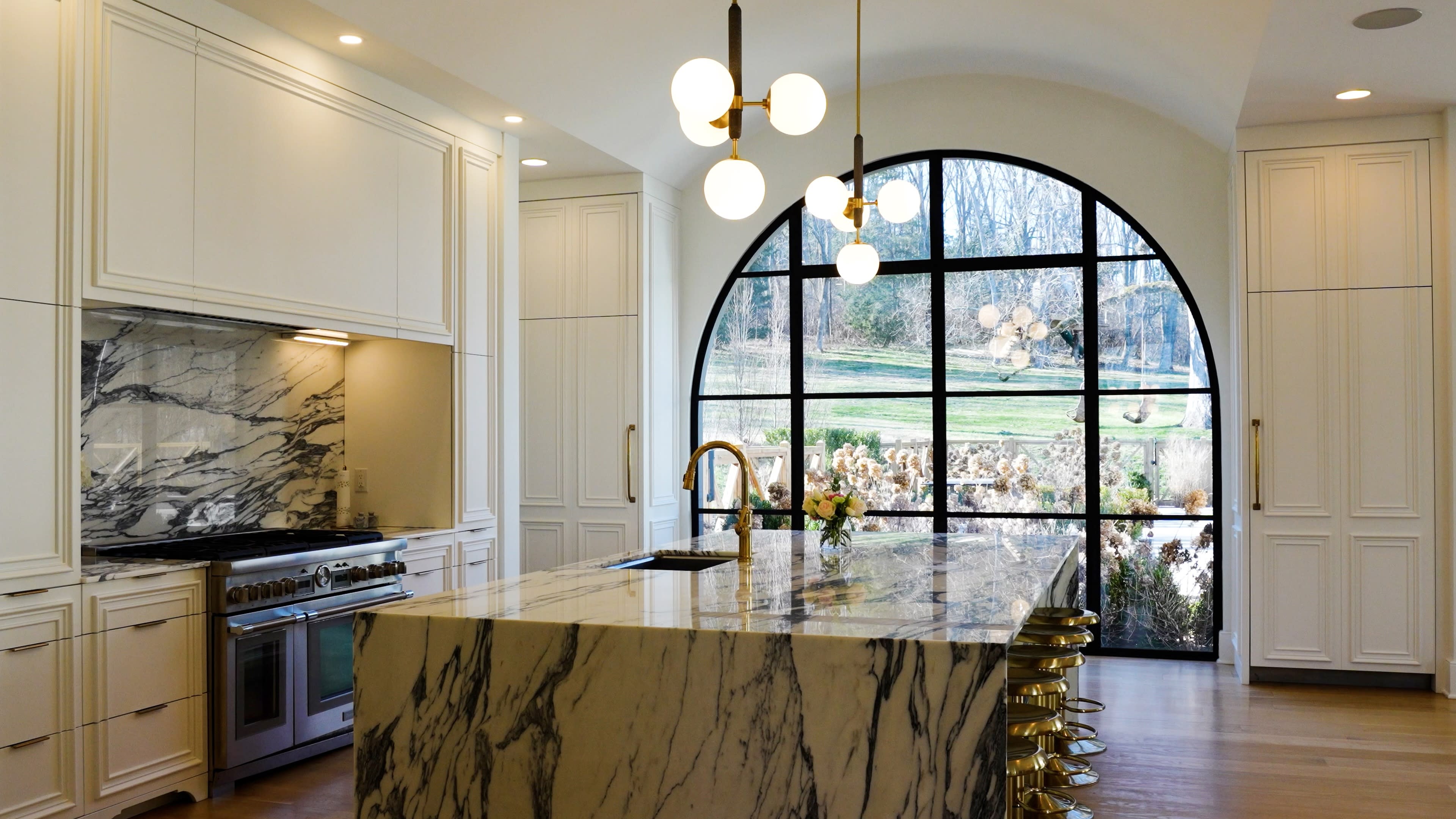 Spacious kitchen with a large marble island, sleek white cabinets, and modern pendant lights. A large arched window offers a scenic garden view.