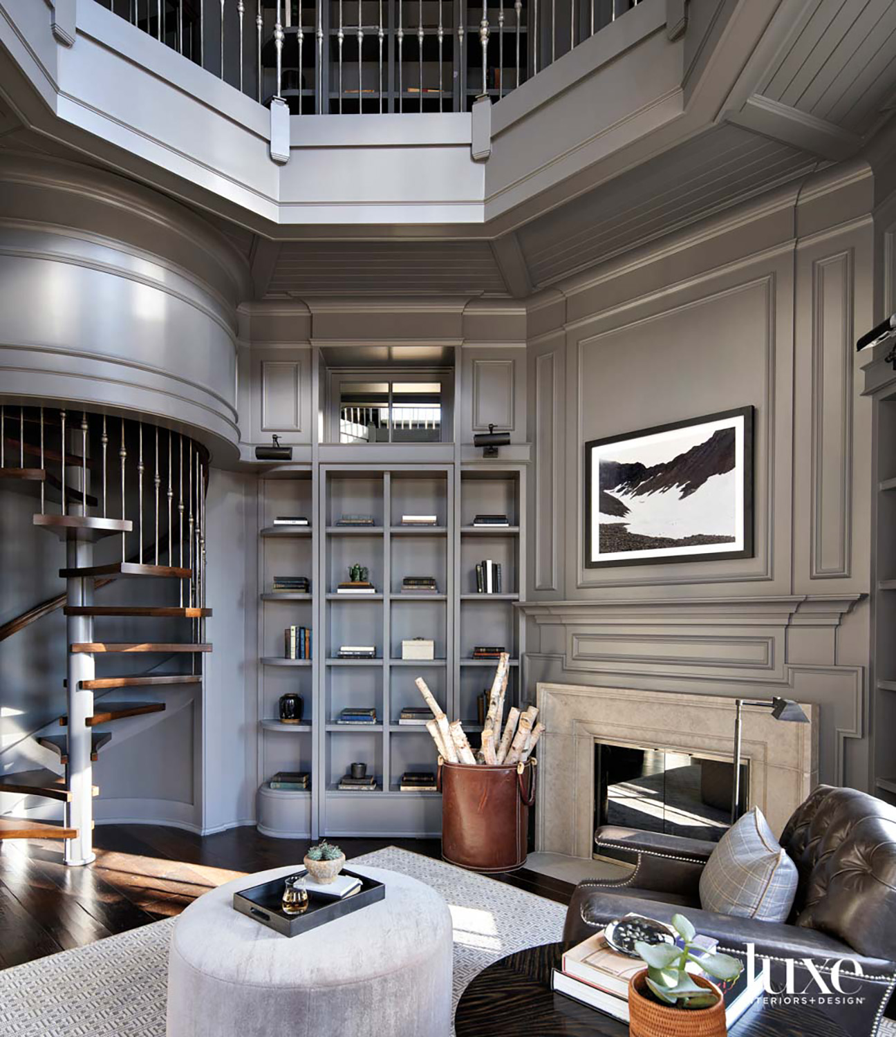 A gray double-height home library with a fireplace and a large leather chair next to a light gray ottoman.