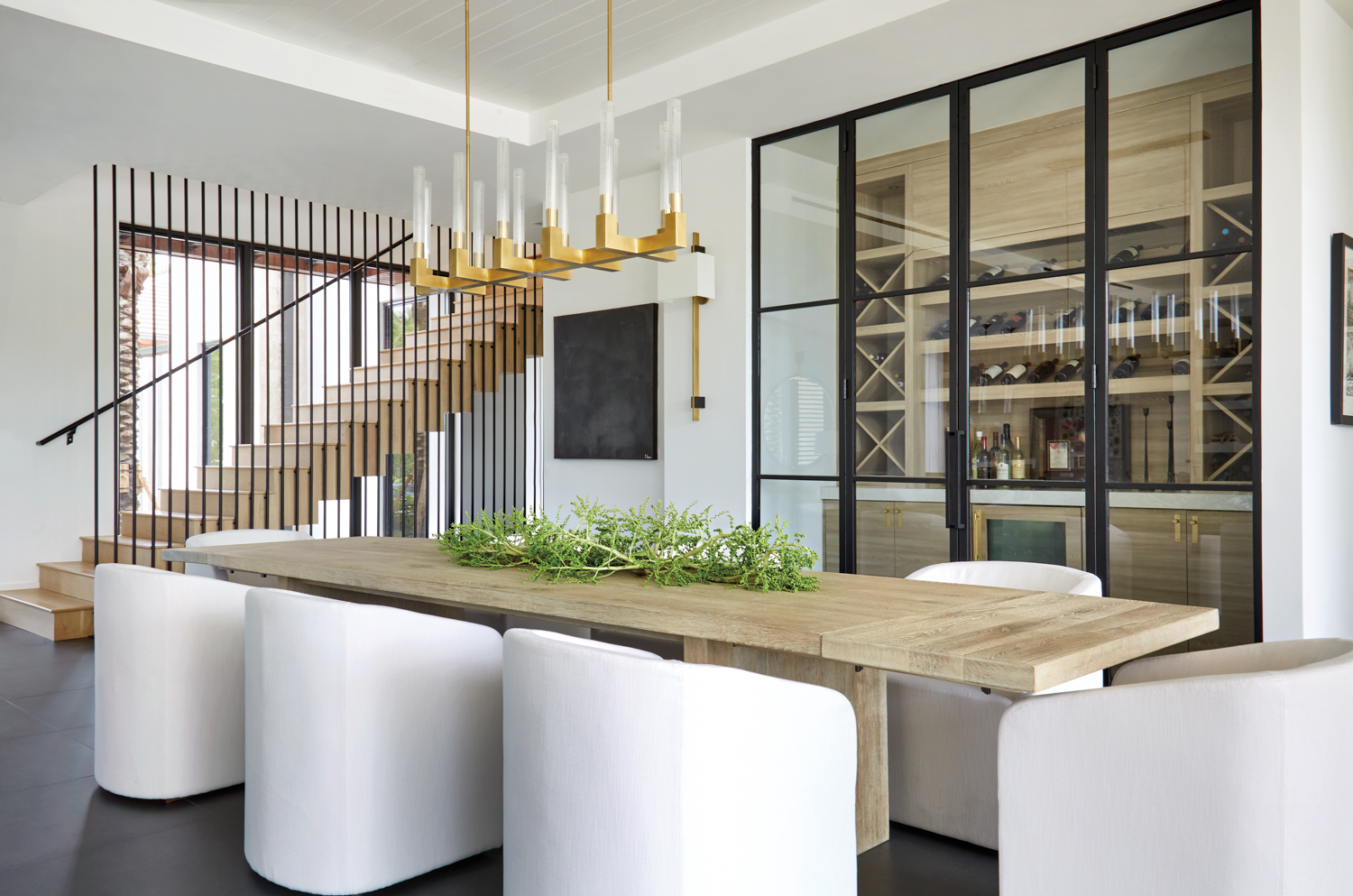 dining area with white chairs, long wood table, wine room, gold chandelier and staircase with iron bars