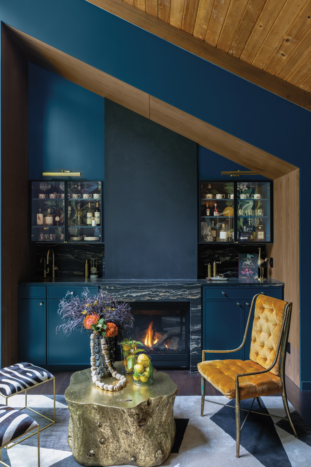 Home wet bar with navy cabinetry and fireplace with marble surround set behind a yellow-velvet-upholstered chair and gold coffee table.