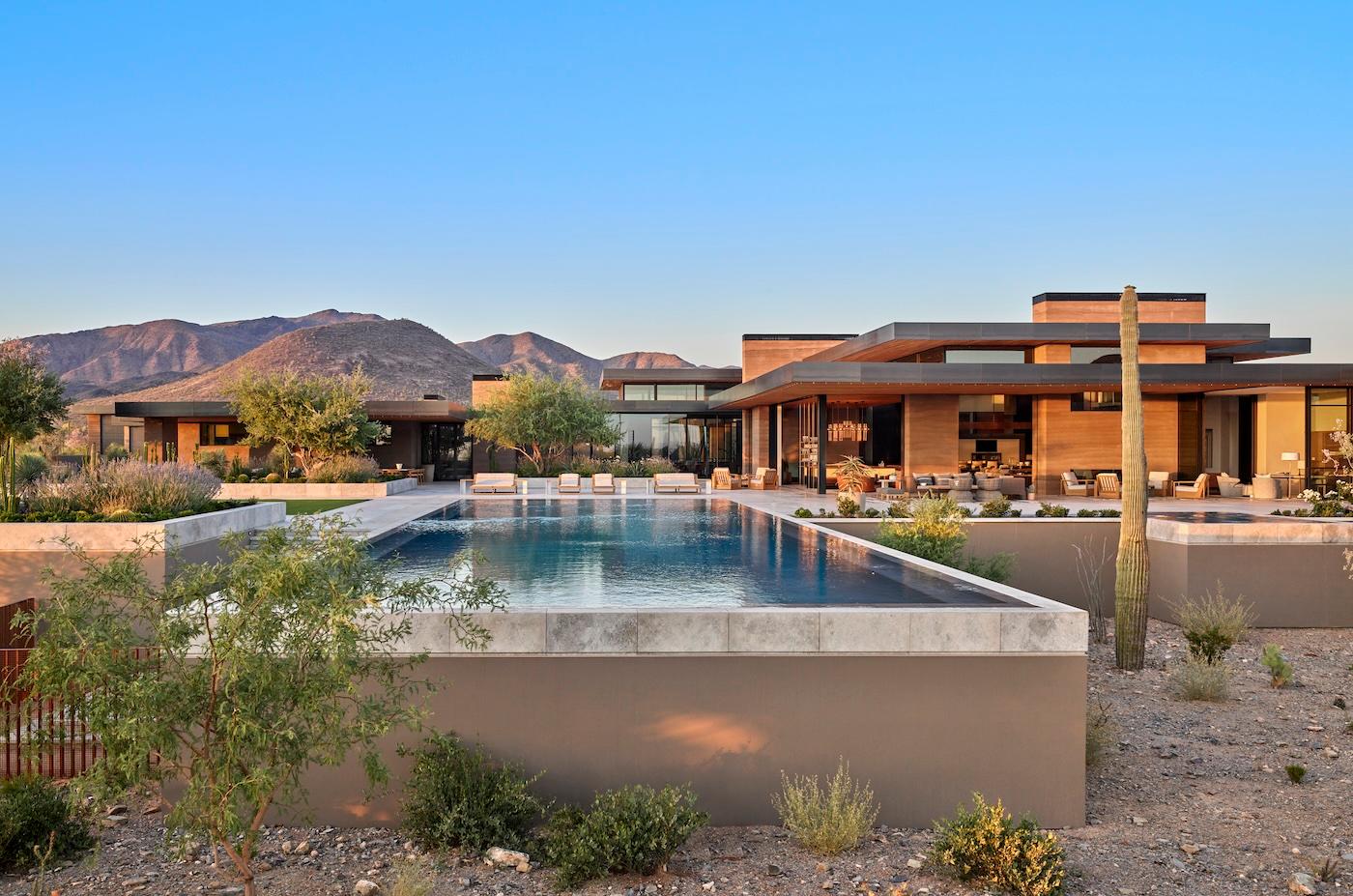 Modern home with a sleek design, featuring a large infinity pool in the foreground. Surrounded by desert landscape and mountains under a clear blue sky.