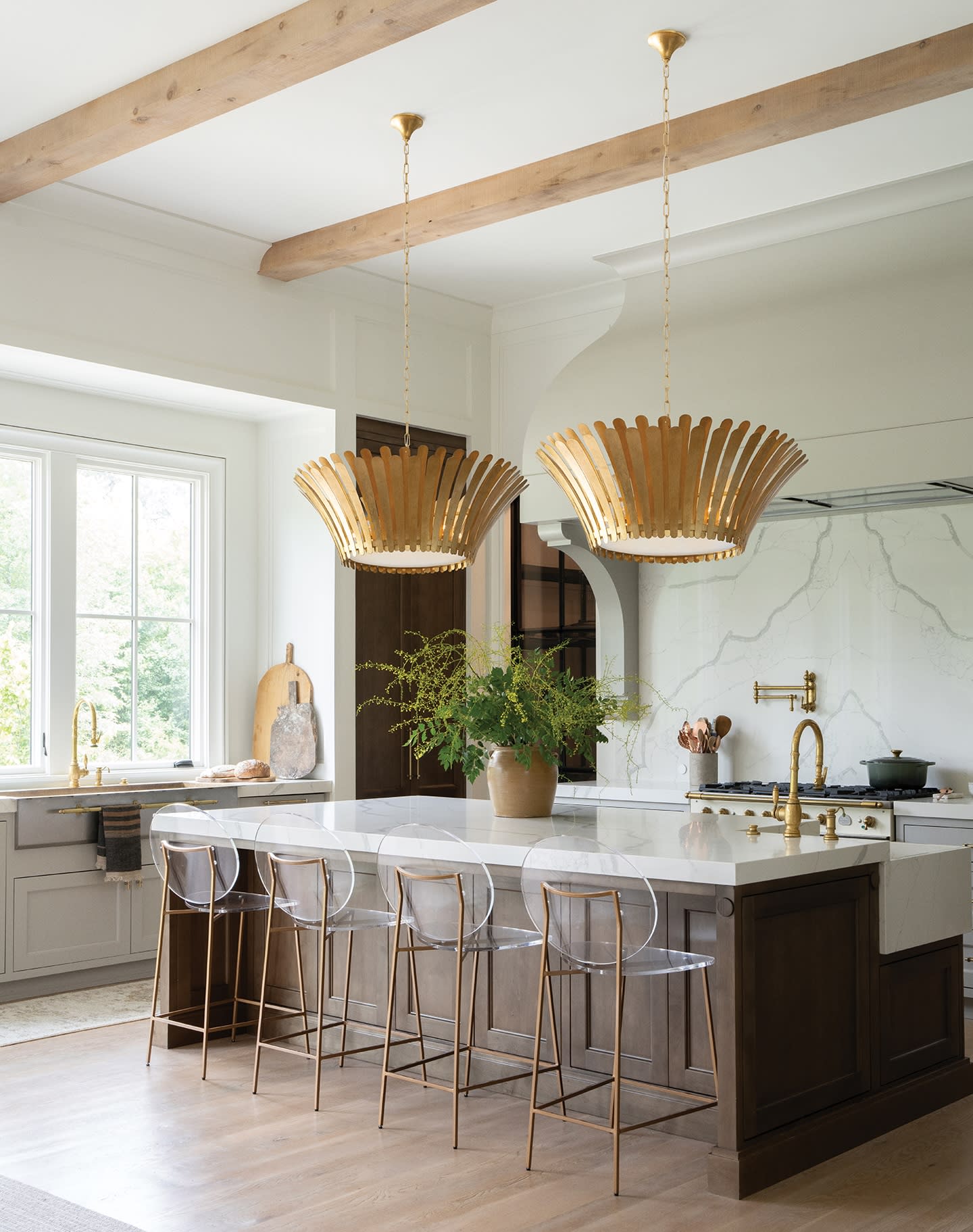 Stylish kitchen with large gold pendant lights, marble island, clear chairs, potted plant, and wood beams. Bright window adds a fresh feel.
