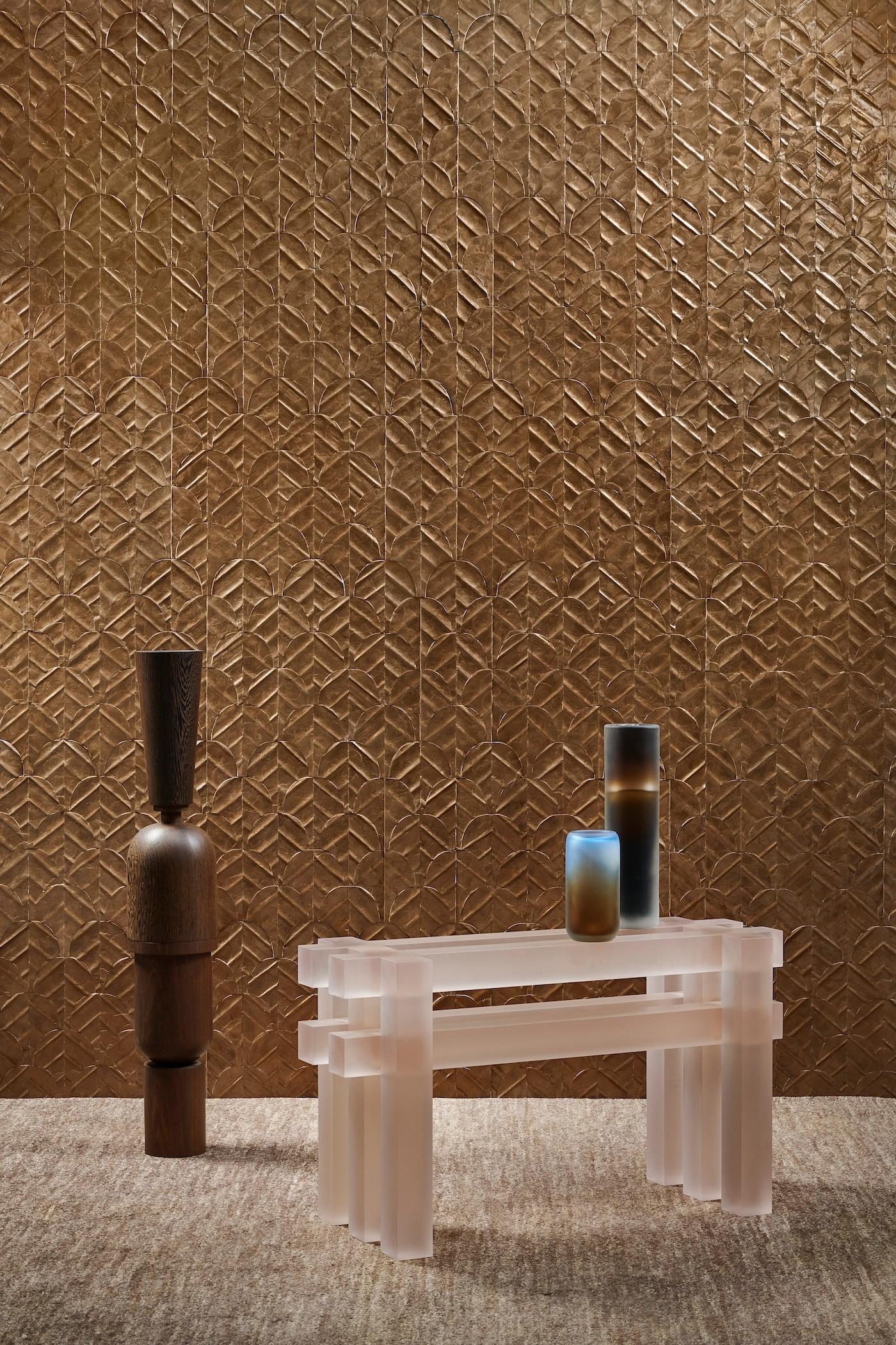 A modern interior scene with a translucent, geometric table on a textured carpet. A wooden vase and two gradient vases sit against a patterned bronze wall, creating a minimalist and elegant atmosphere.