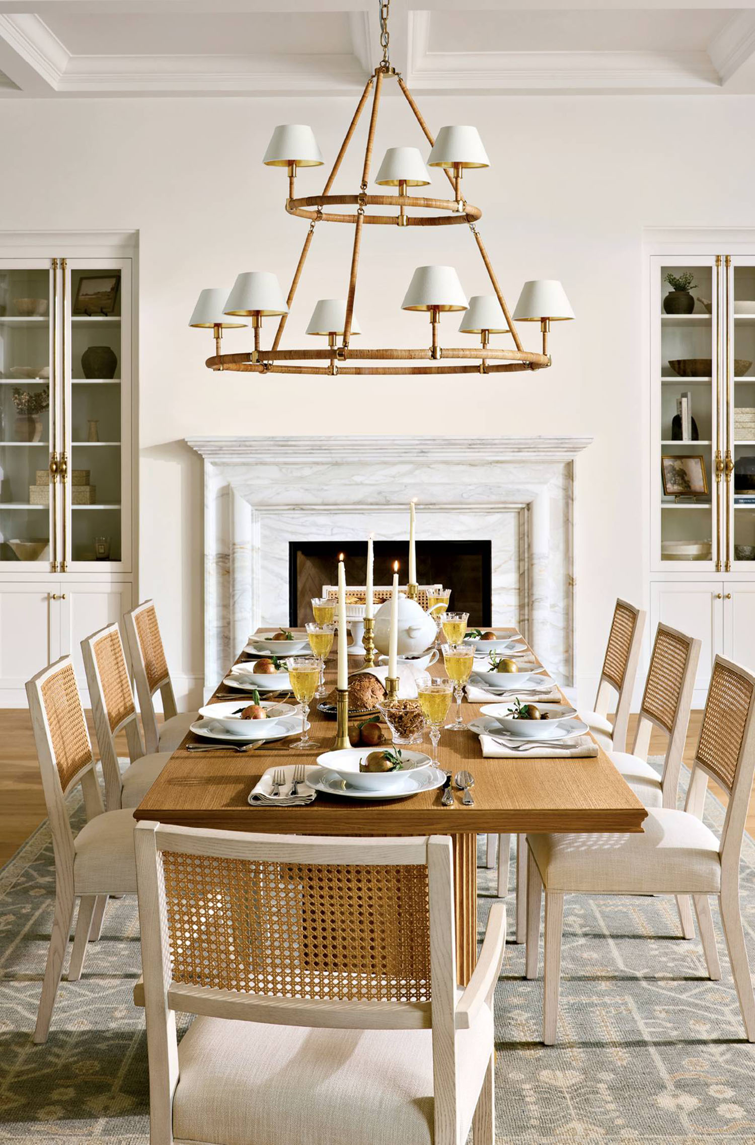 A wood dining table is lined with white chairs with wicker backs, sitting under a chandelier. 
