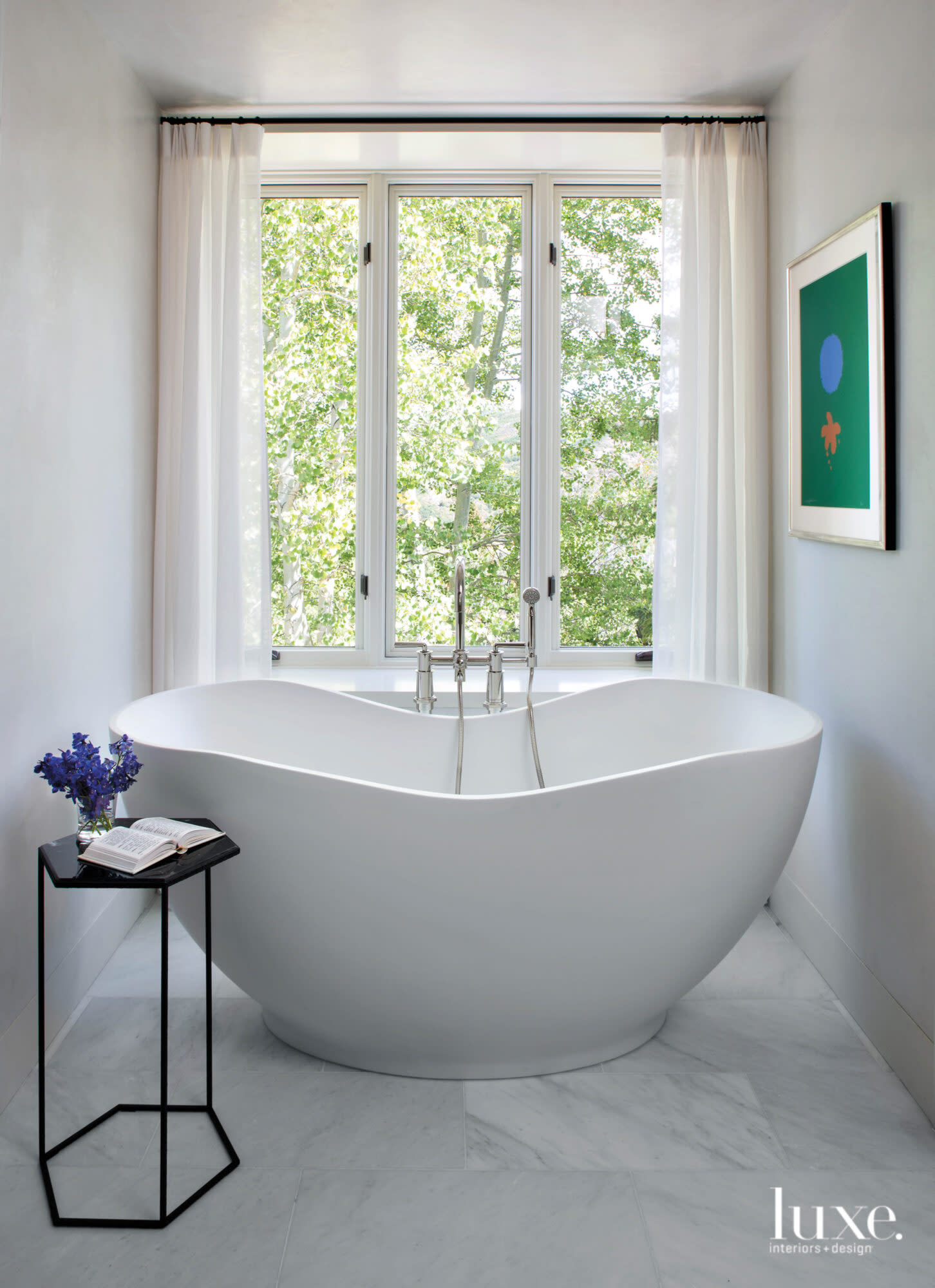 A curvy tub sits in front of a window overlooking a green forest.
