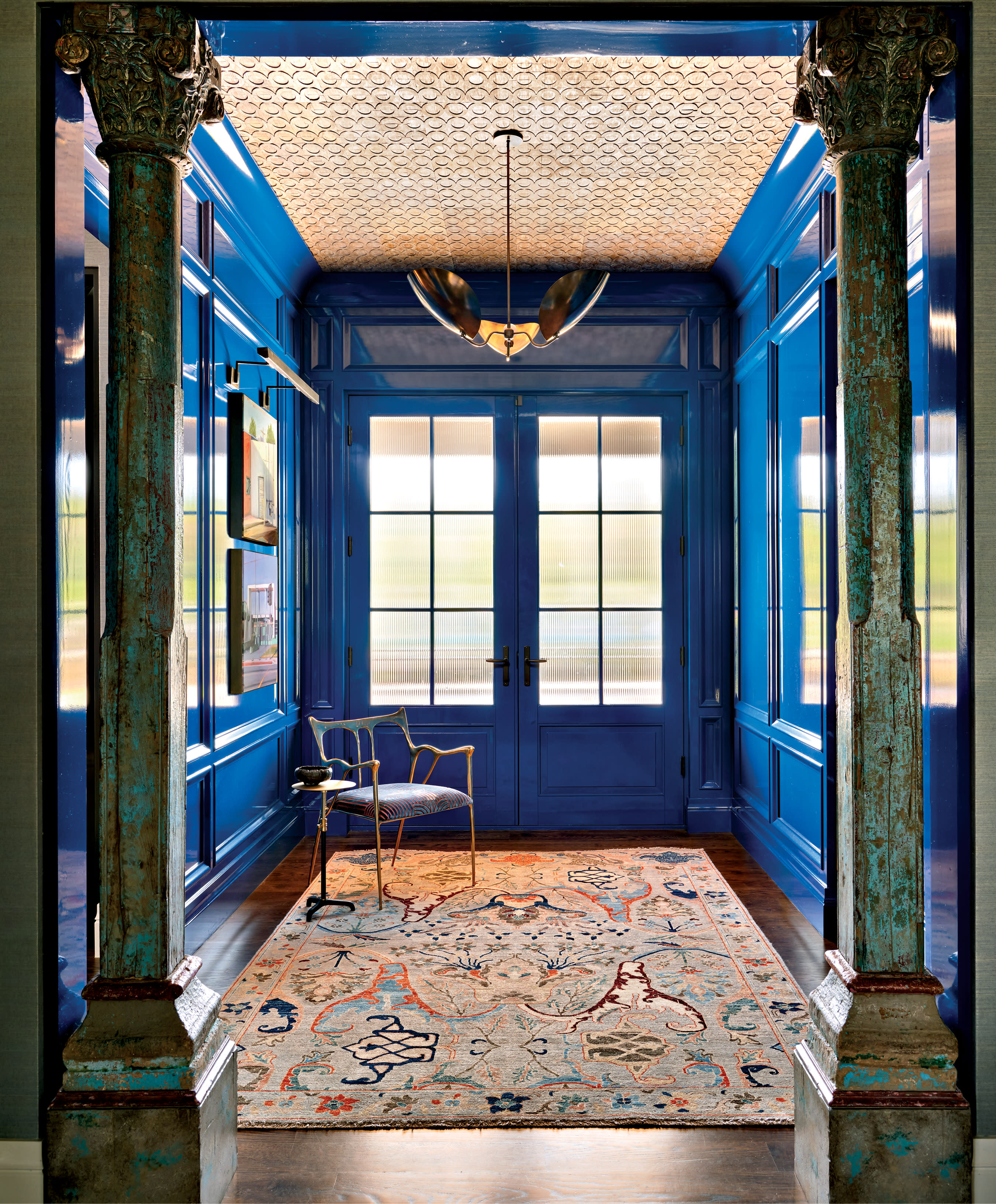Bright blue hallway with ornate columns, a patterned rug, and a single chair. Large windows reflect natural light, creating an inviting and elegant space.